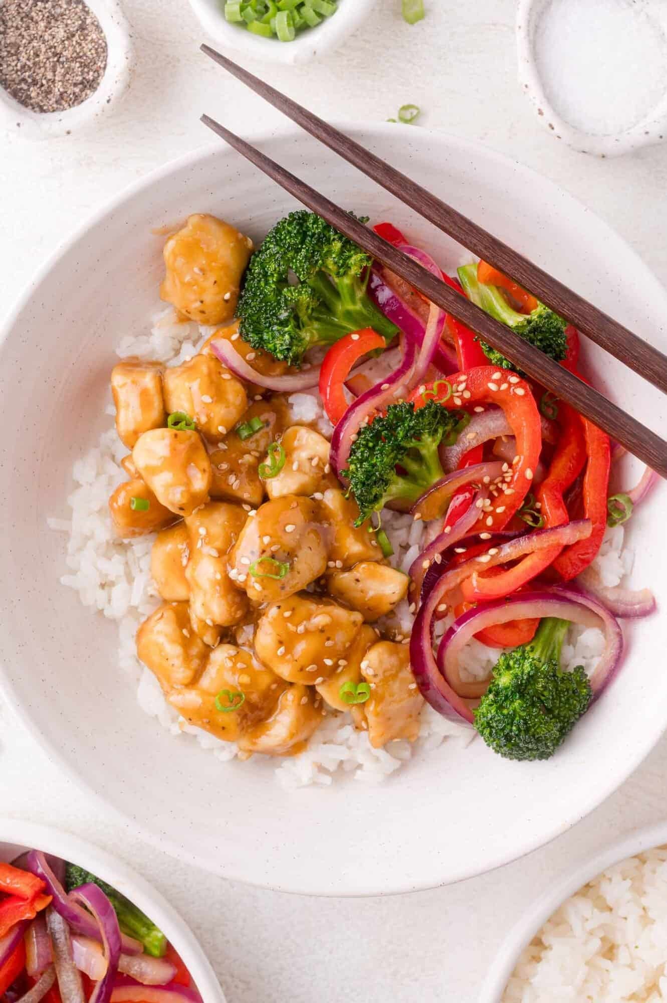 A bowl of white rice topped with sesame chicken, broccoli, red bell pepper, and red onion, with chopsticks resting on the bowl.