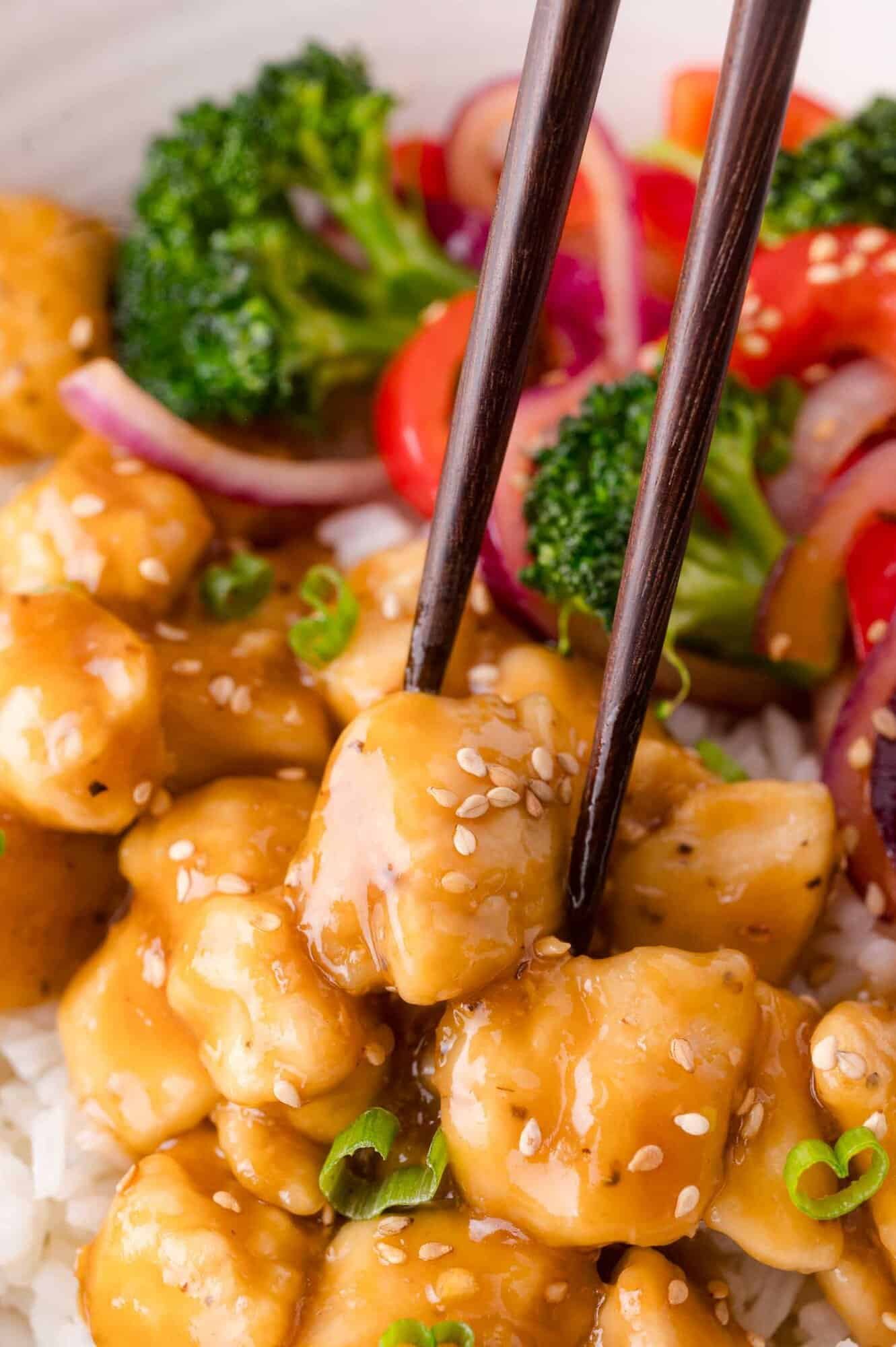 Chopsticks holding a piece of teriyaki chicken over white rice, with broccoli and sliced vegetables in the background.