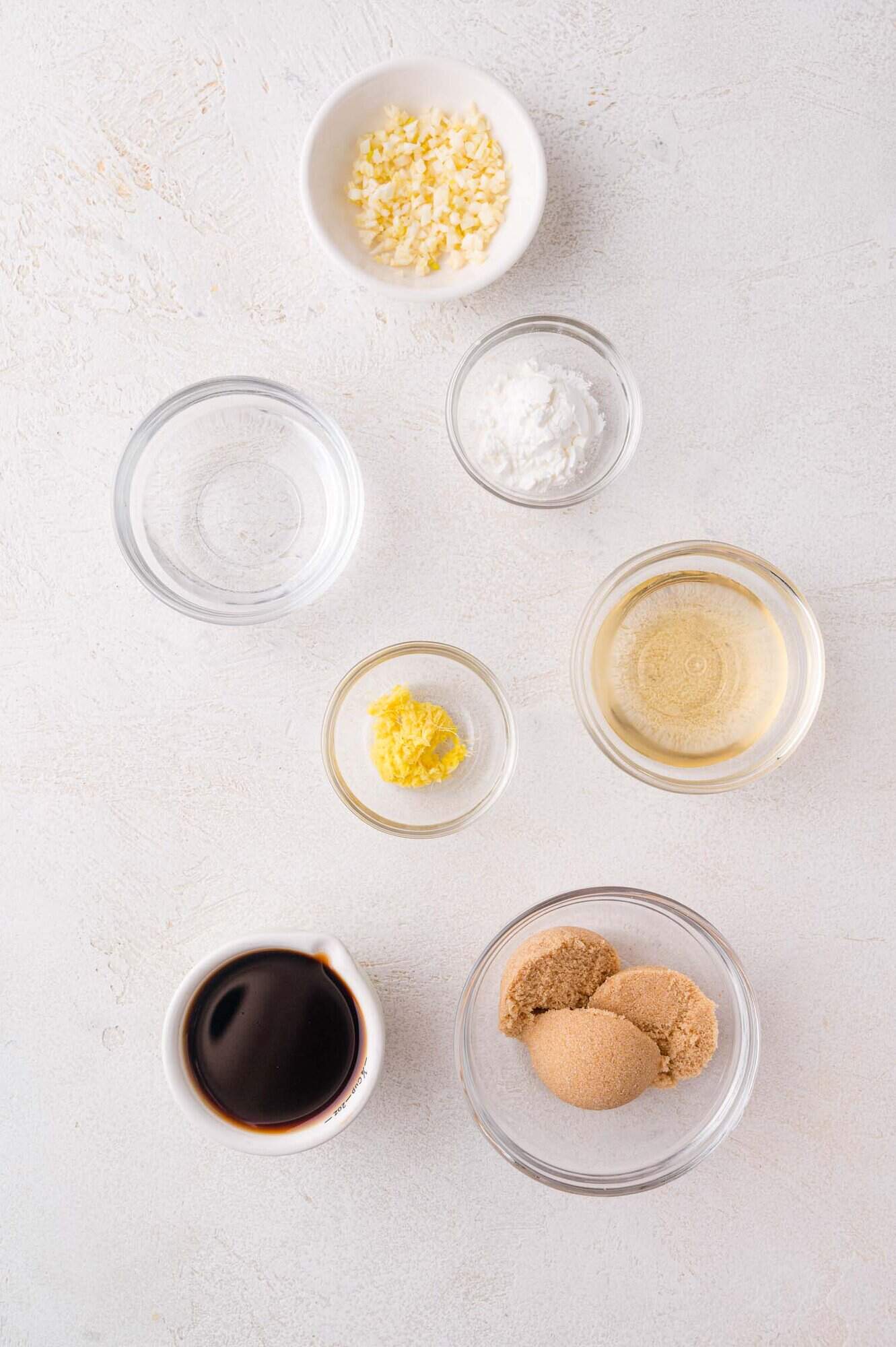 Seven small glass and ceramic bowls containing minced garlic, cornstarch, water, grated ginger, rice vinegar, soy sauce, and brown sugar arranged on a white surface.