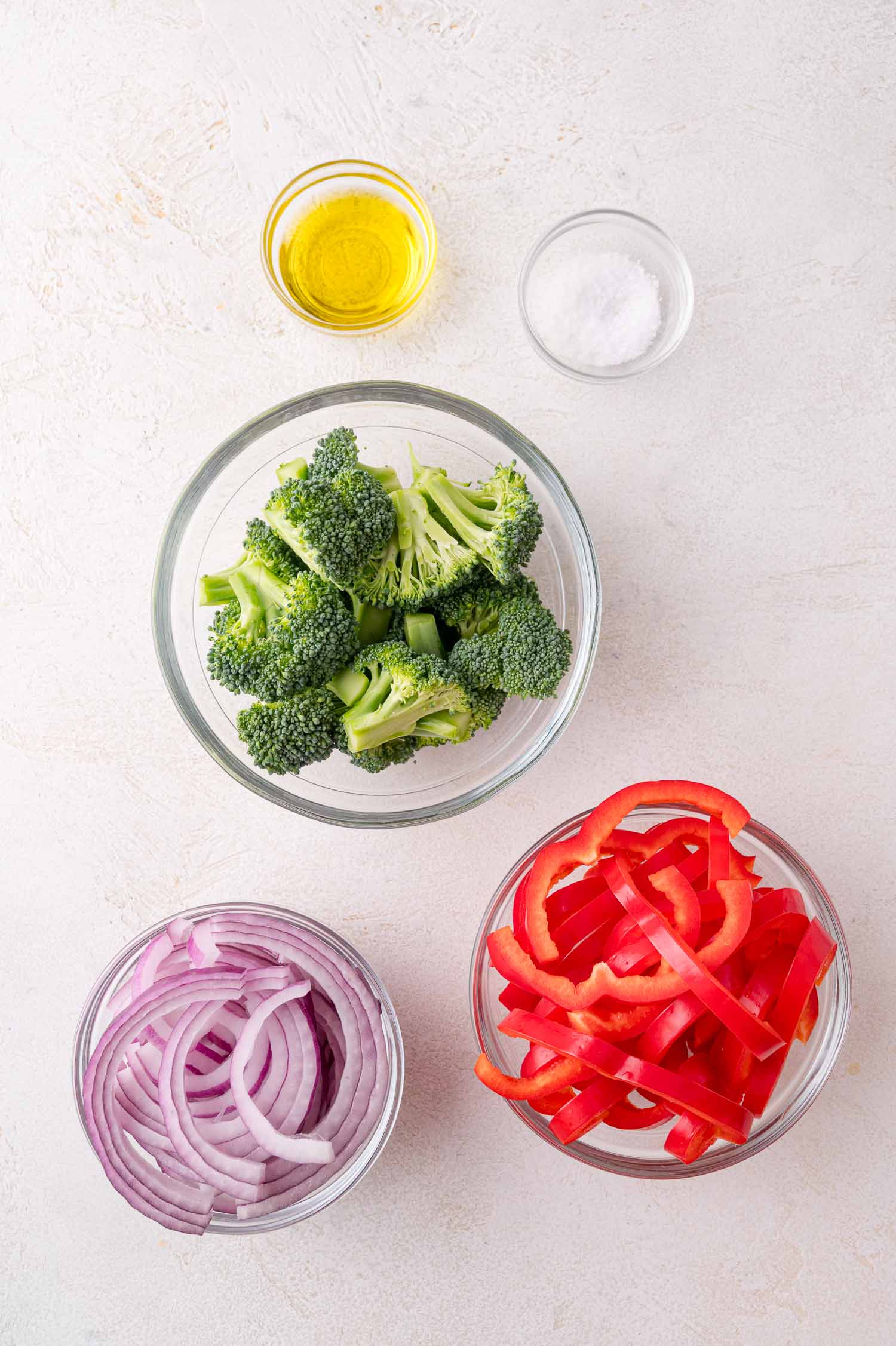 Three bowls containing broccoli florets, sliced red bell pepper, and sliced red onion, with small dishes of olive oil and salt on a light surface.