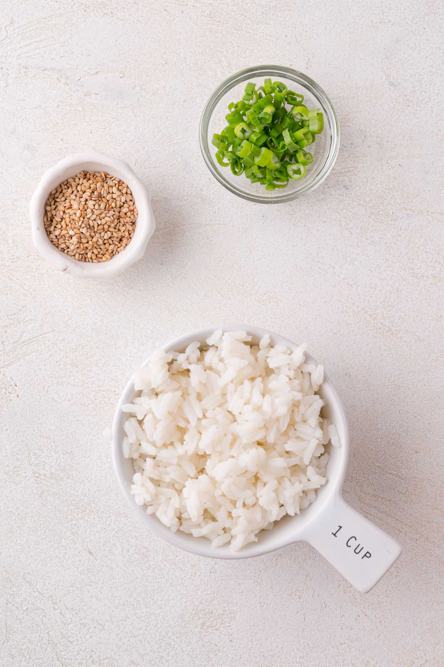 A cup of cooked white rice, a small bowl of sliced green onions, and a small bowl of sesame seeds on a light surface.