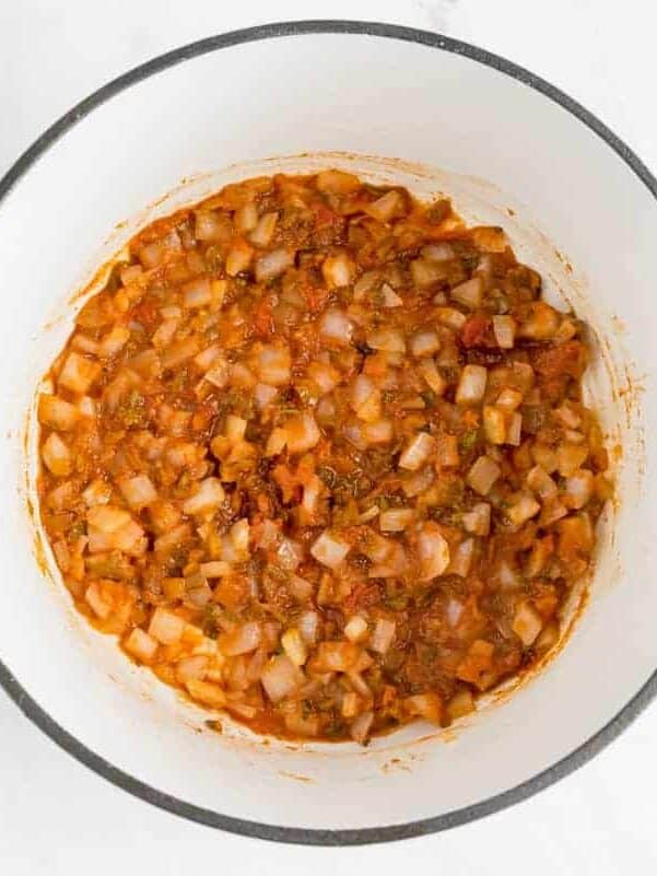 Chopped onions and tomatoes cooking in a white pot with blue handles, placed on a white surface next to a light blue cloth.