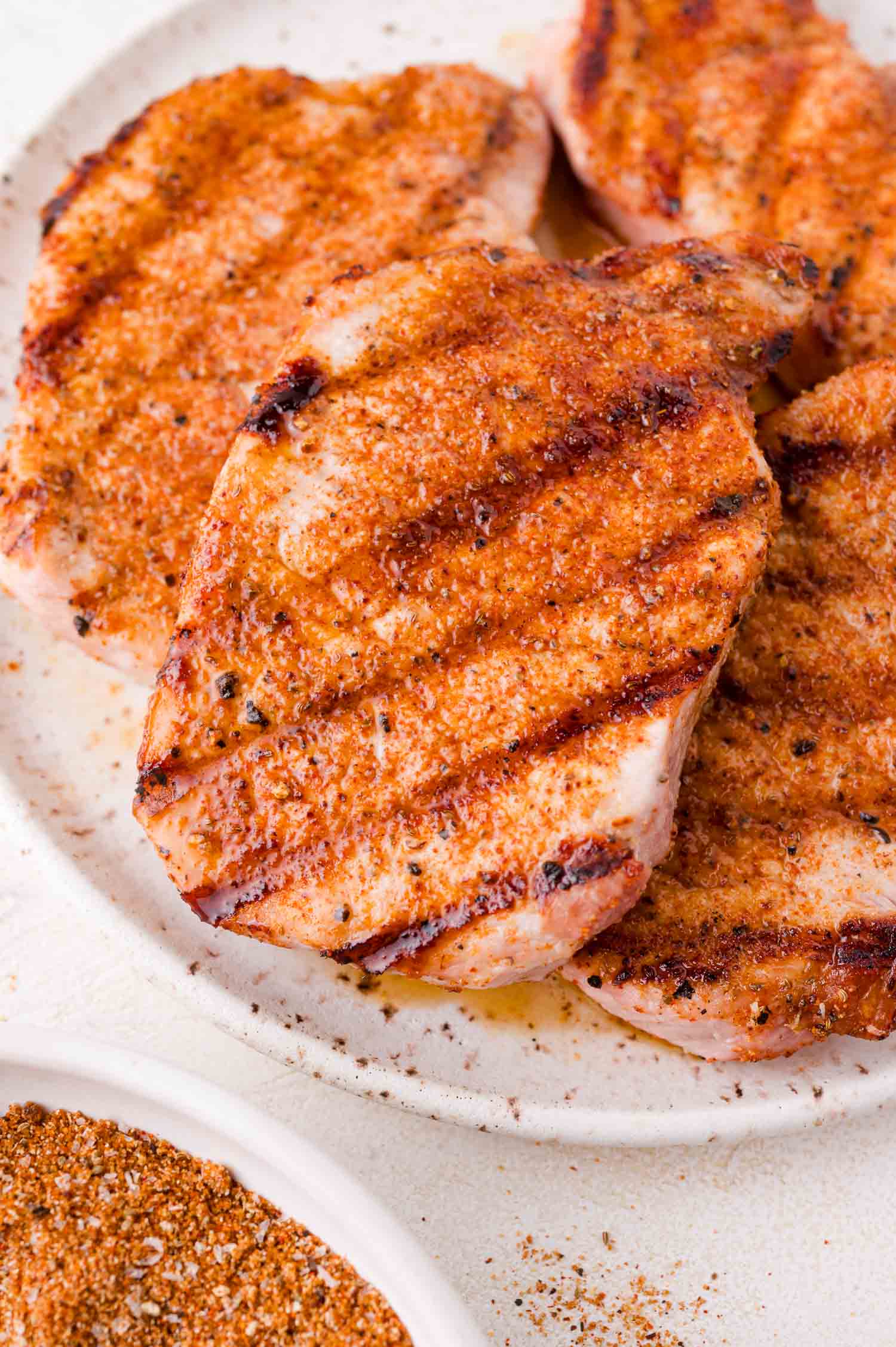 Three grilled pork chops with a seasoned spice rub on a white plate, showing grill marks and a bowl of spice mix nearby.