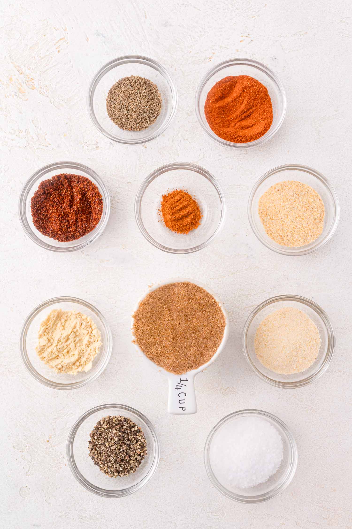 Ten small glass bowls and one measuring cup, each filled with different spices and seasonings, are arranged in a grid pattern on a white surface.