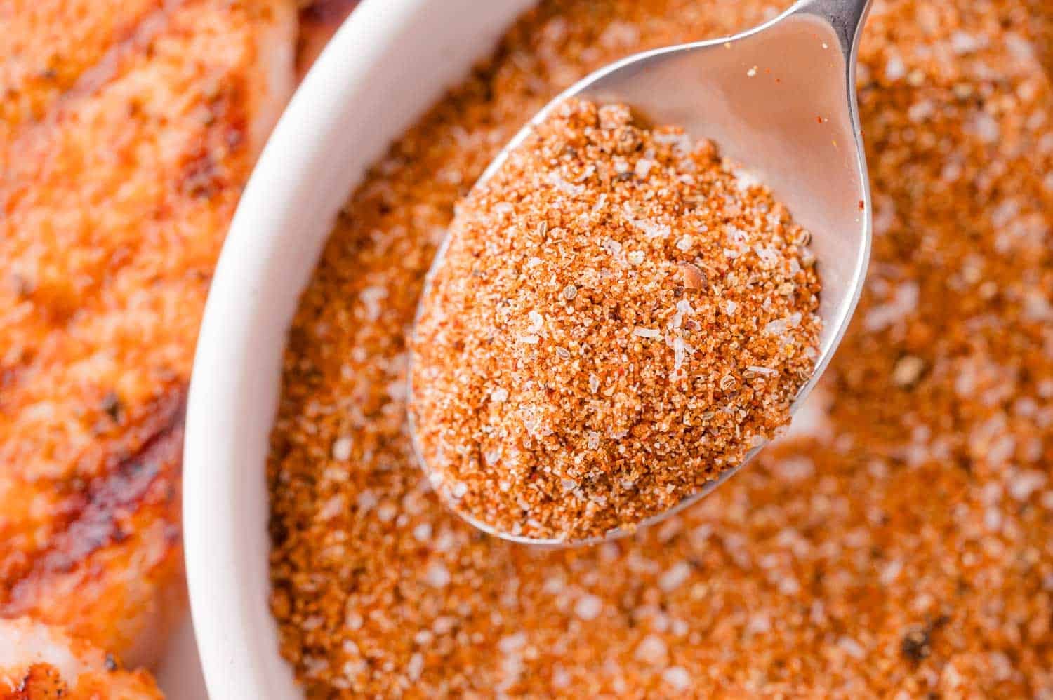 A close-up of a spoonful of dry seasoning mix held over a white bowl filled with the same spice blend.