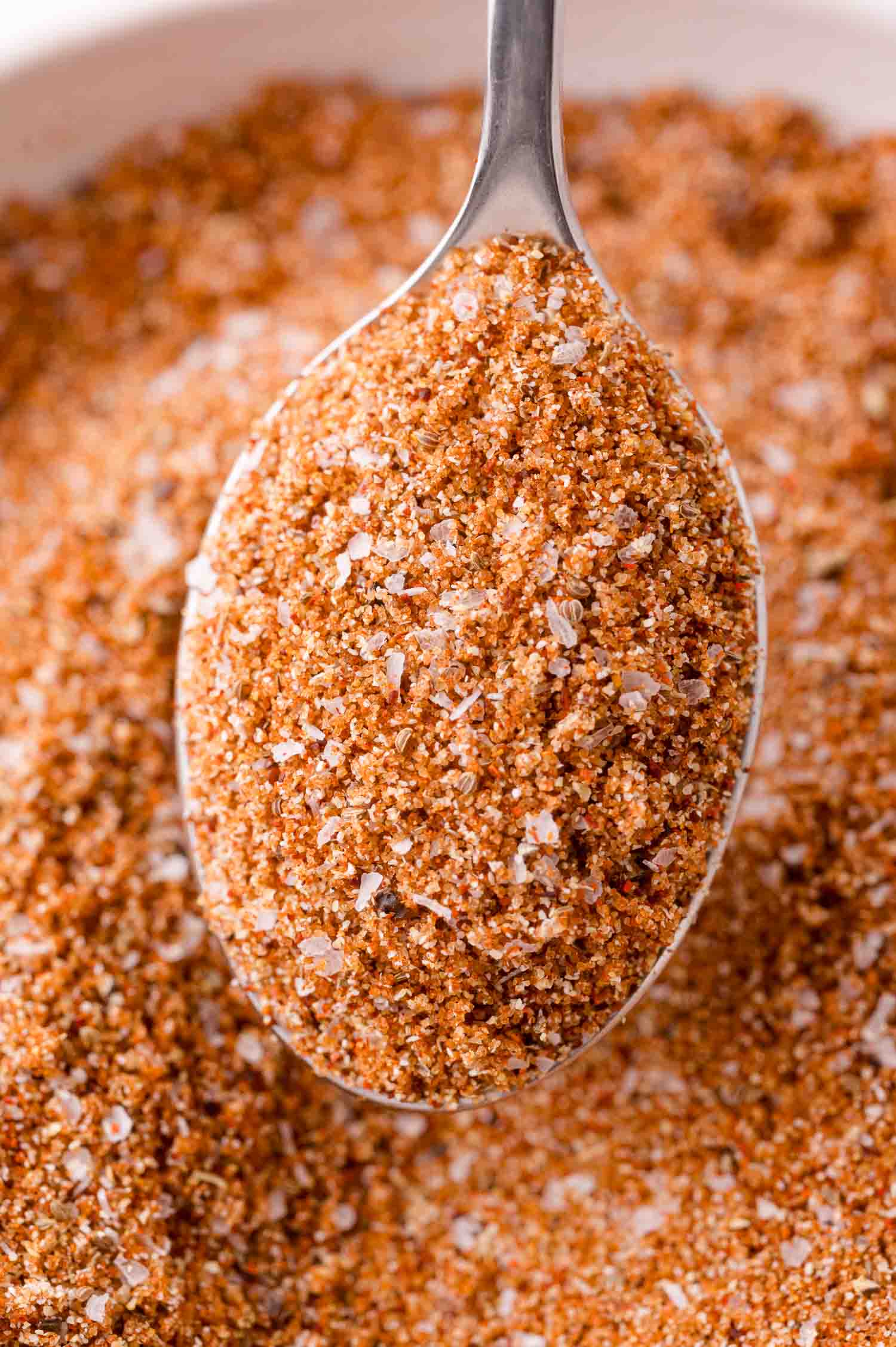 A close-up of a spoonful of coarse, mixed bbq rub, featuring visible salt granules, held over a bowl filled with the same mixture.