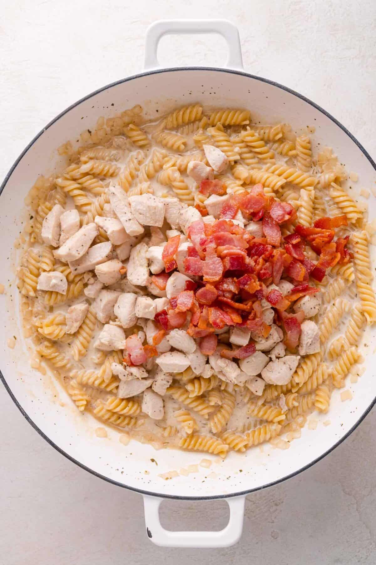 A white pot contains cooked rotini pasta, diced chicken, and chopped bacon, all mixed in a creamy sauce.
