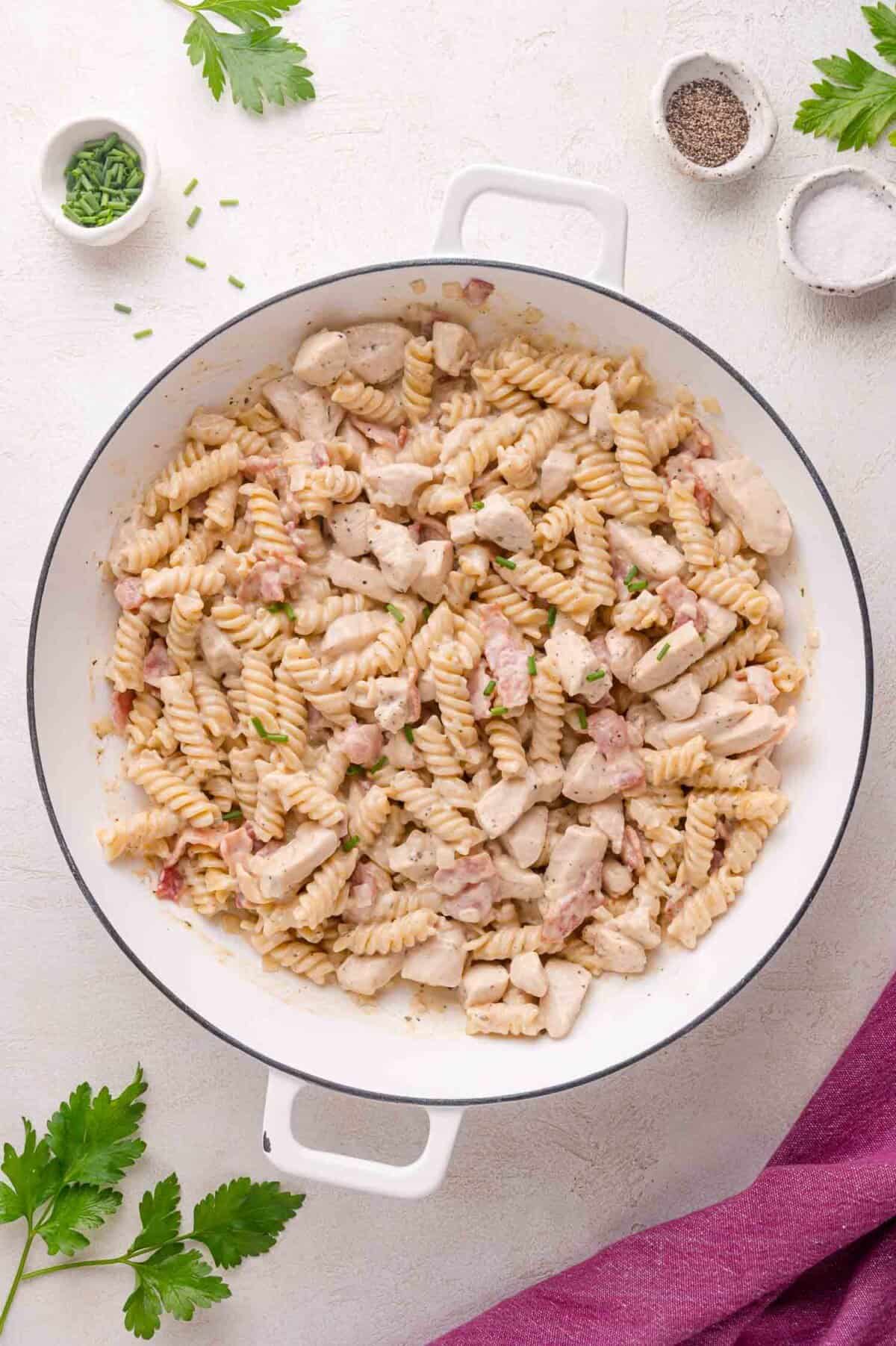 A white pan filled with cooked rotini pasta, diced chicken, and small pieces of bacon, garnished with chopped herbs. Small bowls of pepper and herbs are nearby.