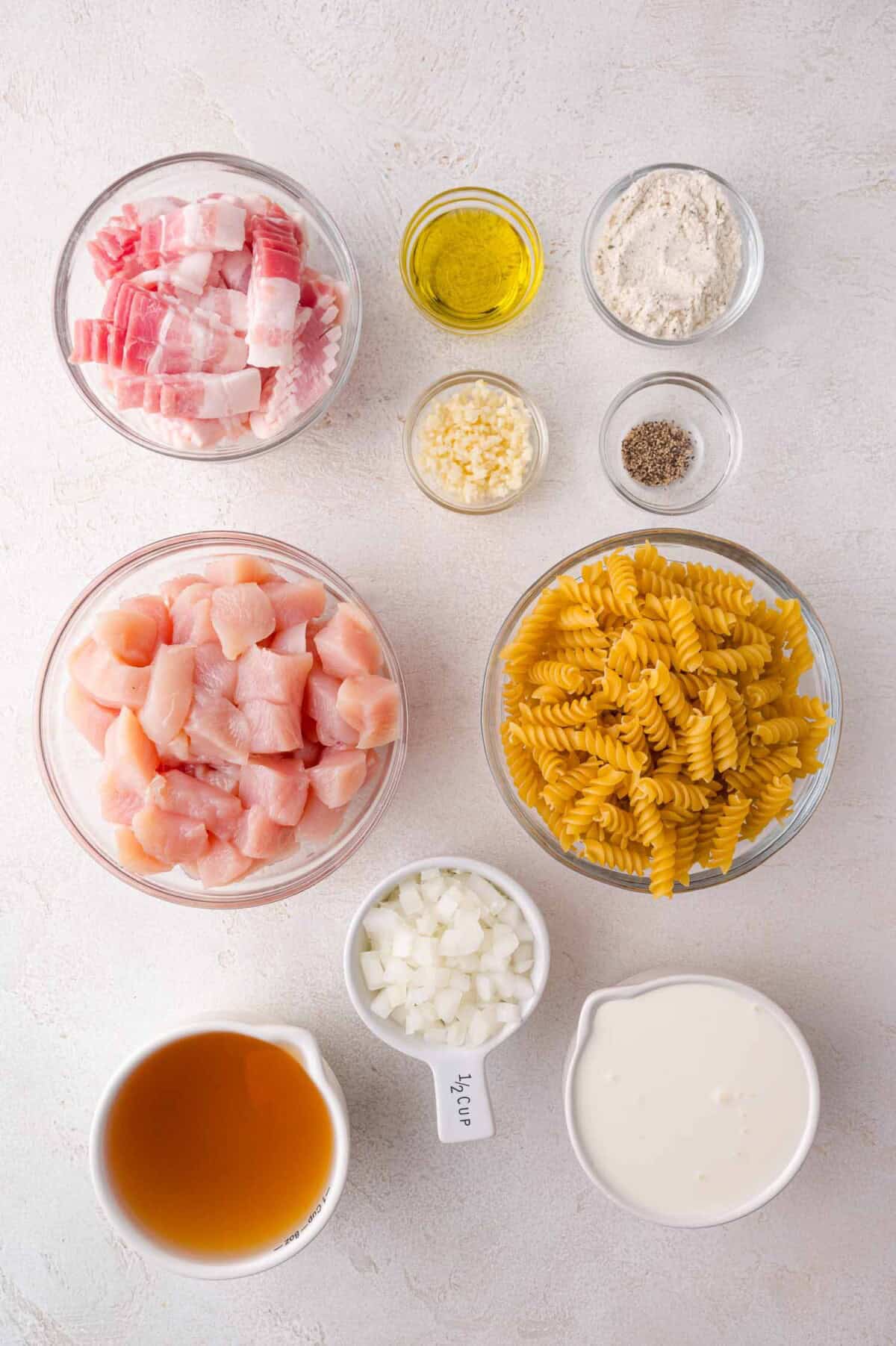 Bowls and containers with raw chicken, bacon pieces, dry rotini pasta, cream, diced onion, flour, oil, minced garlic, pepper, and broth on a white surface.