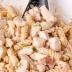 Close-up of creamy rotini pasta mixed with diced chicken, bacon pieces, and garnished with chopped chives. A black serving spoon is partially visible.
