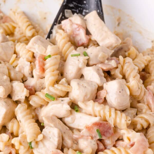 Close-up of creamy rotini pasta mixed with diced chicken, bacon pieces, and garnished with chopped chives. A black serving spoon is partially visible.