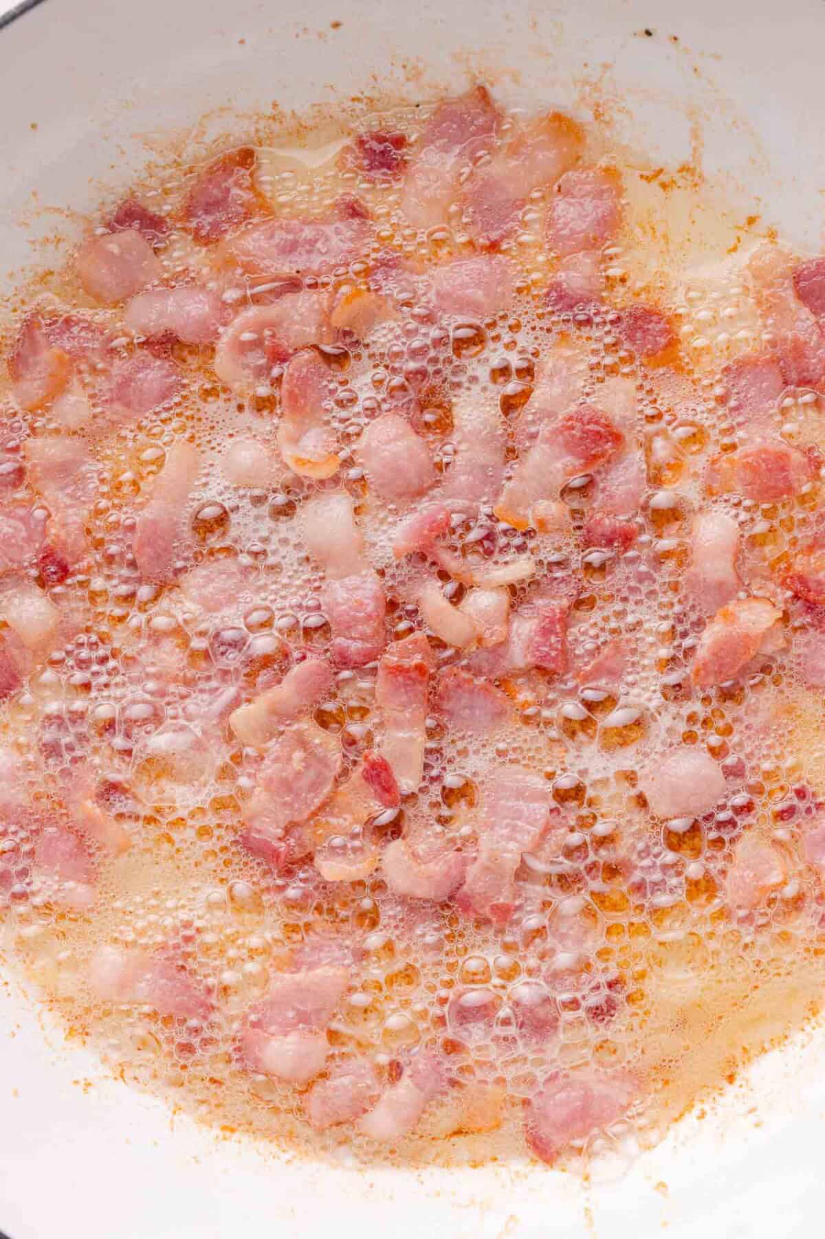 Small pieces of bacon are sizzling and bubbling in hot oil in a frying pan.