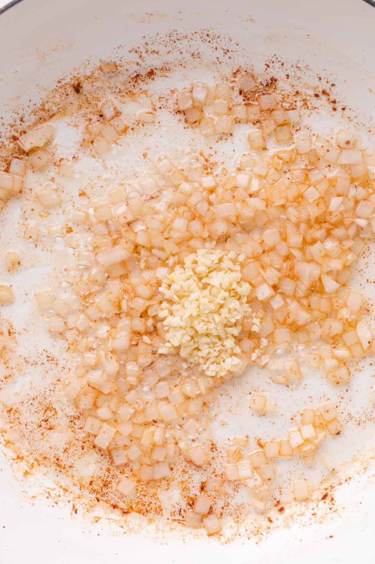 Diced onions are sautéed in a pan with browned bits, with a mound of minced garlic added in the center.