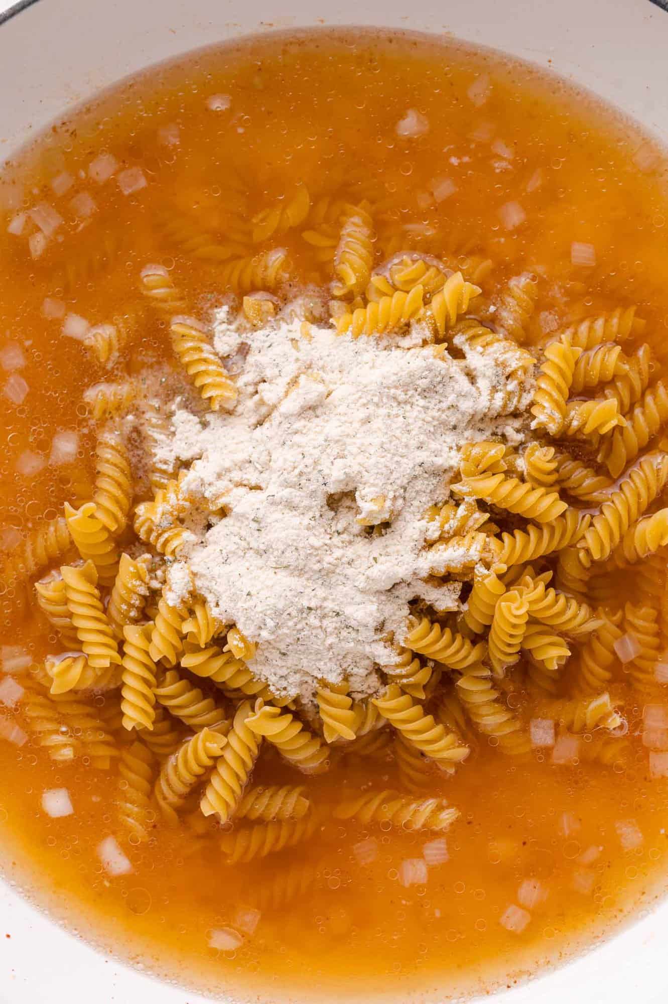 Uncooked rotini pasta, a mound of ranch seasoning, and diced onions in broth inside a white pot.