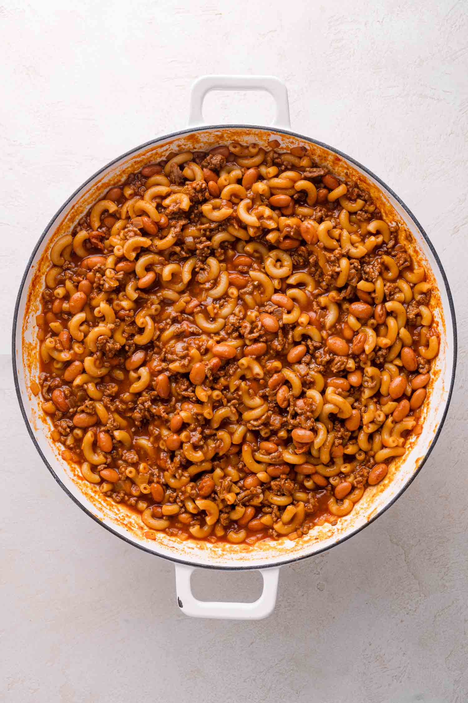 A white pot filled with cooked macaroni, ground meat, beans, and tomato sauce, mixed together.