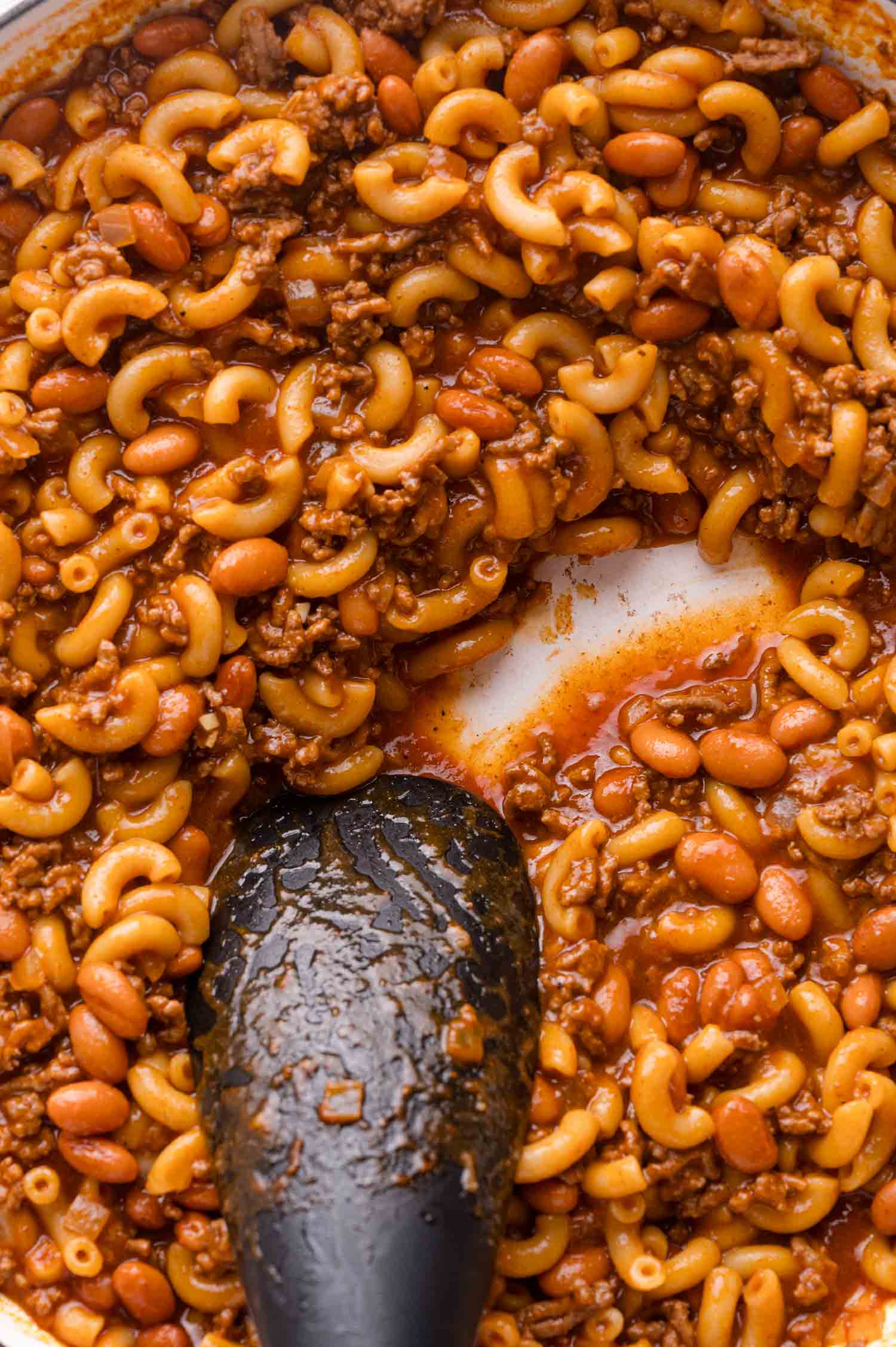 A close-up of cooked macaroni, ground beef, and beans in a tomato-based sauce, with a black spoon stirring the mixture in a pot.