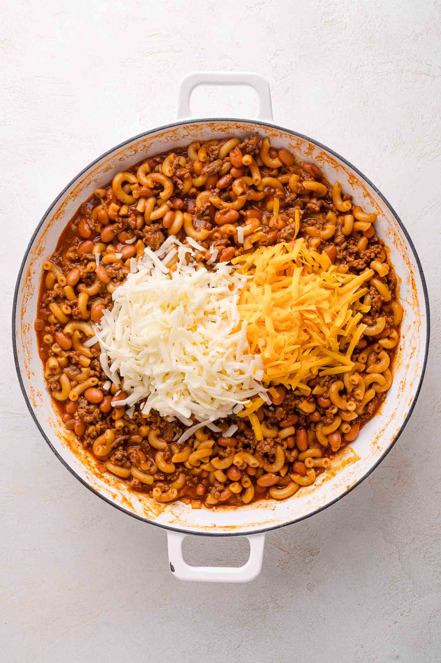 A white pot filled with cooked macaroni, ground meat, beans, tomato sauce, and topped with shredded white and yellow cheese.