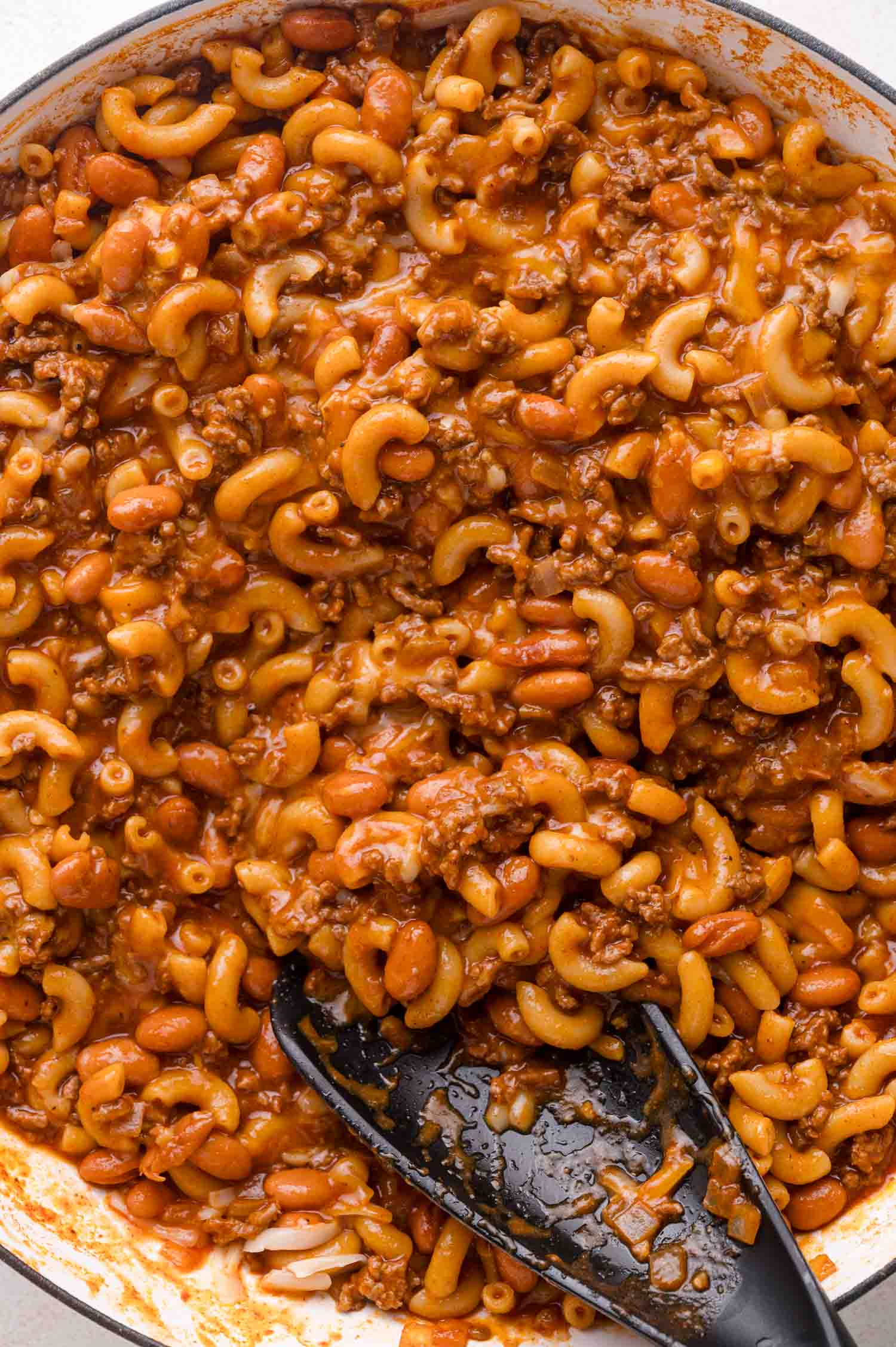 Close-up of cooked chili mac with elbow macaroni, ground beef, beans, and tomato sauce in a pan, with a black serving spoon.