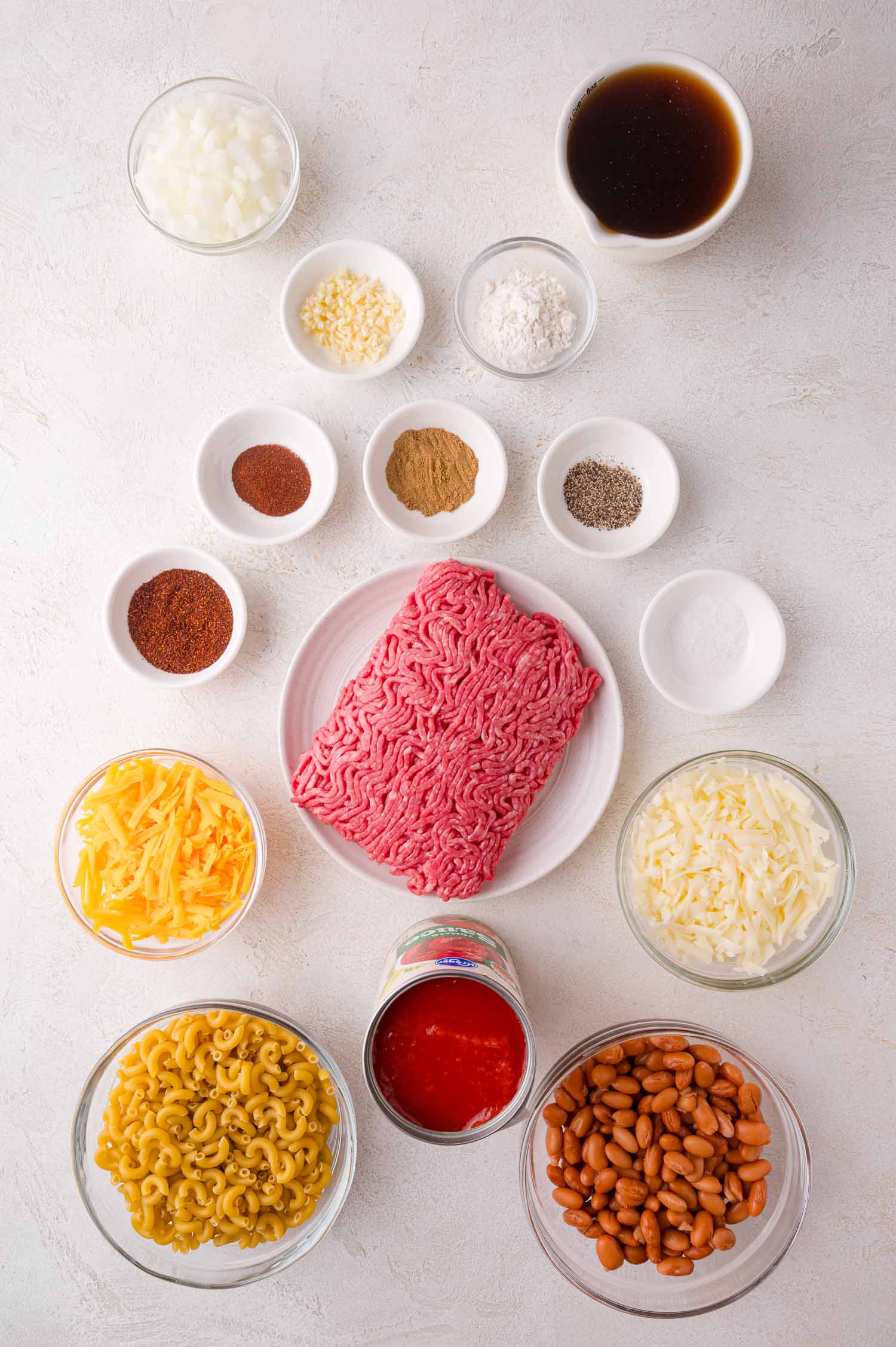 Ingredients for chili mac arranged in bowls on a white surface, including ground beef, beans, pasta, cheeses, tomato, spices, onion, garlic, and broth.