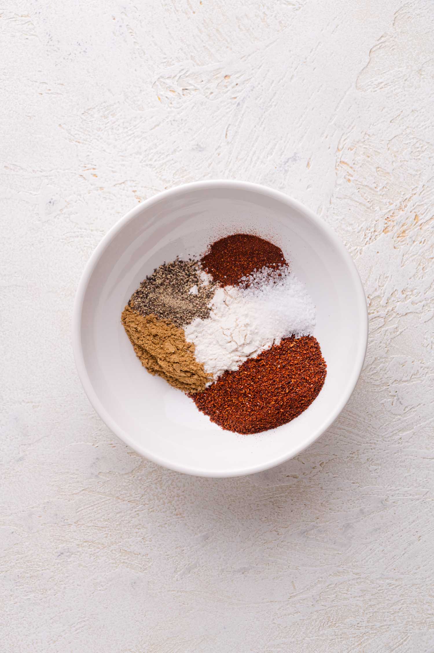 A white bowl containing five different ground spices and seasonings arranged in separate piles on a light textured surface.