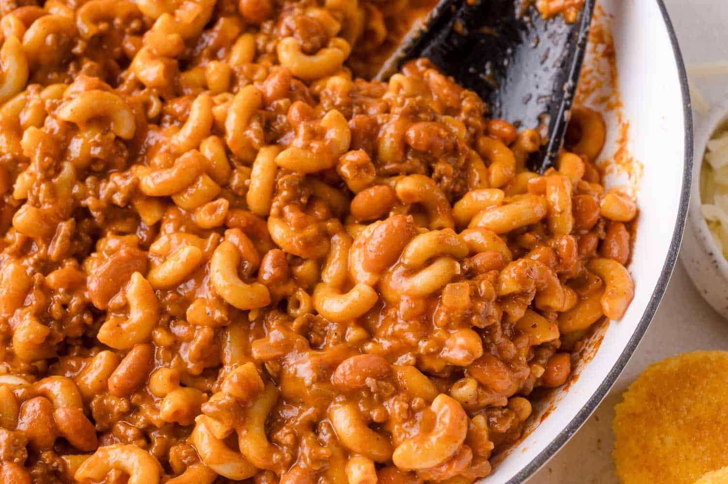 Close-up of chili mac in a pan with elbow macaroni, beans, ground meat, and tomato sauce, with a black serving spoon and a cornbread muffin on the side.