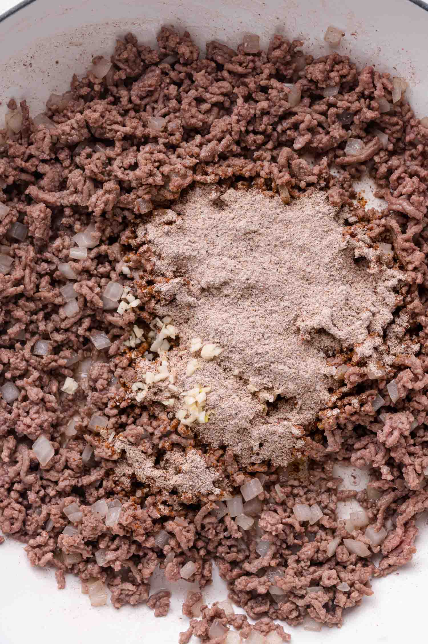 Ground beef and diced onions cooking in a pan with a pile of seasoning mix and minced garlic added to the center.