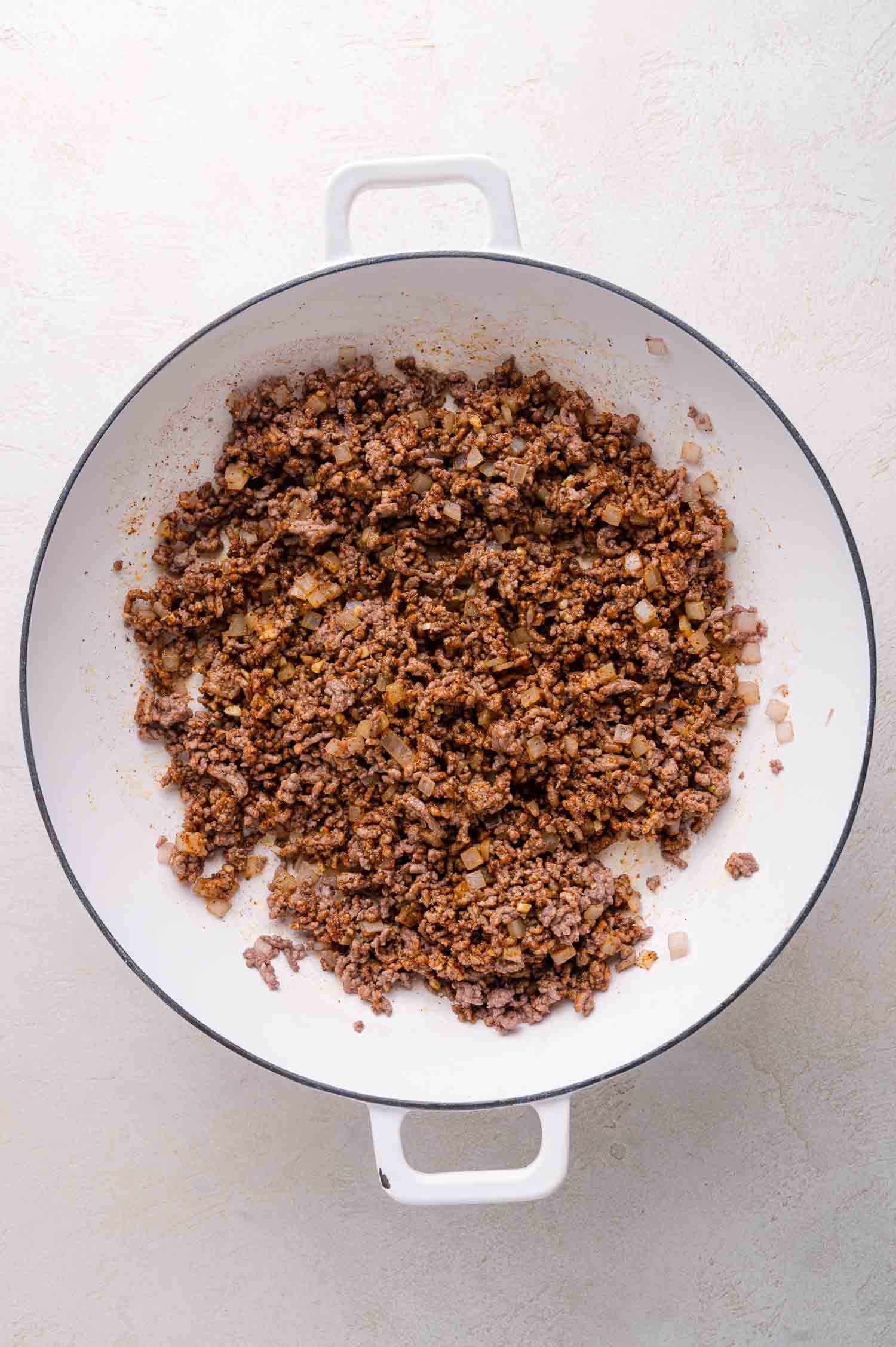 Cooked ground beef with diced onions and seasoning in a white pan on a light surface.