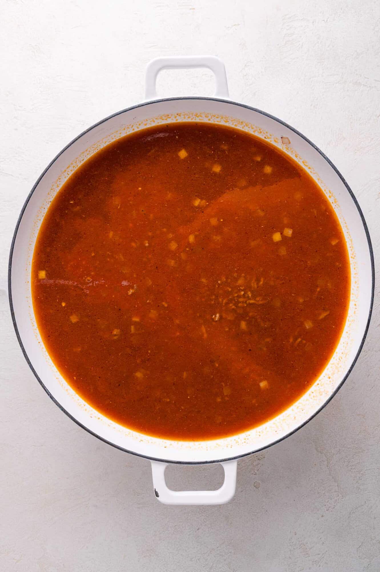 A white pot filled with red broth containing small pieces of vegetables and beef, viewed from above on a light surface.