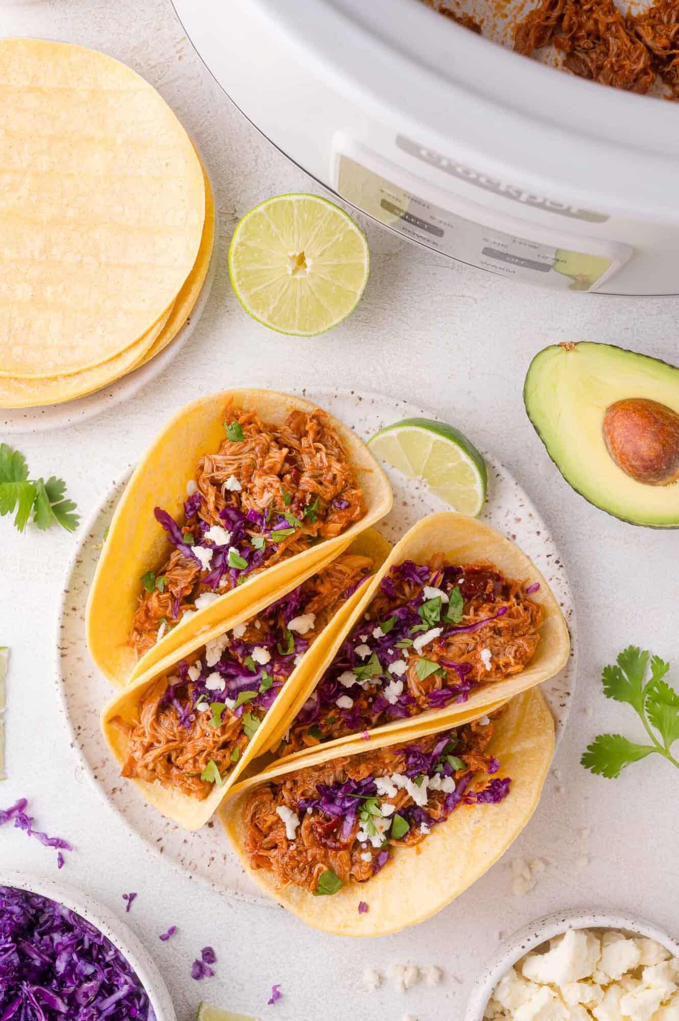 Three shredded chicken tacos in corn tortillas on a plate, garnished with chopped cilantro, crumbled cheese, and shredded purple cabbage. Avocado, lime, and tortillas are nearby.