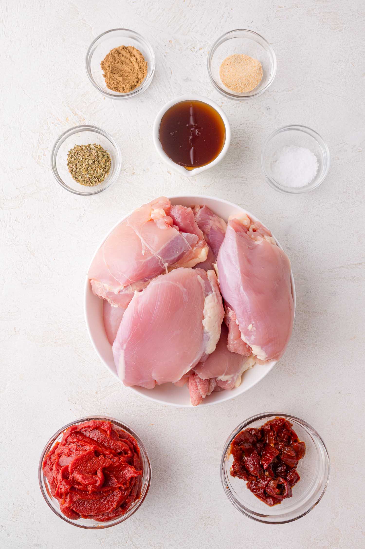 Raw chicken thighs in a bowl surrounded by small bowls of spices, chipotle chiles, and adobo sauce on a white surface.
