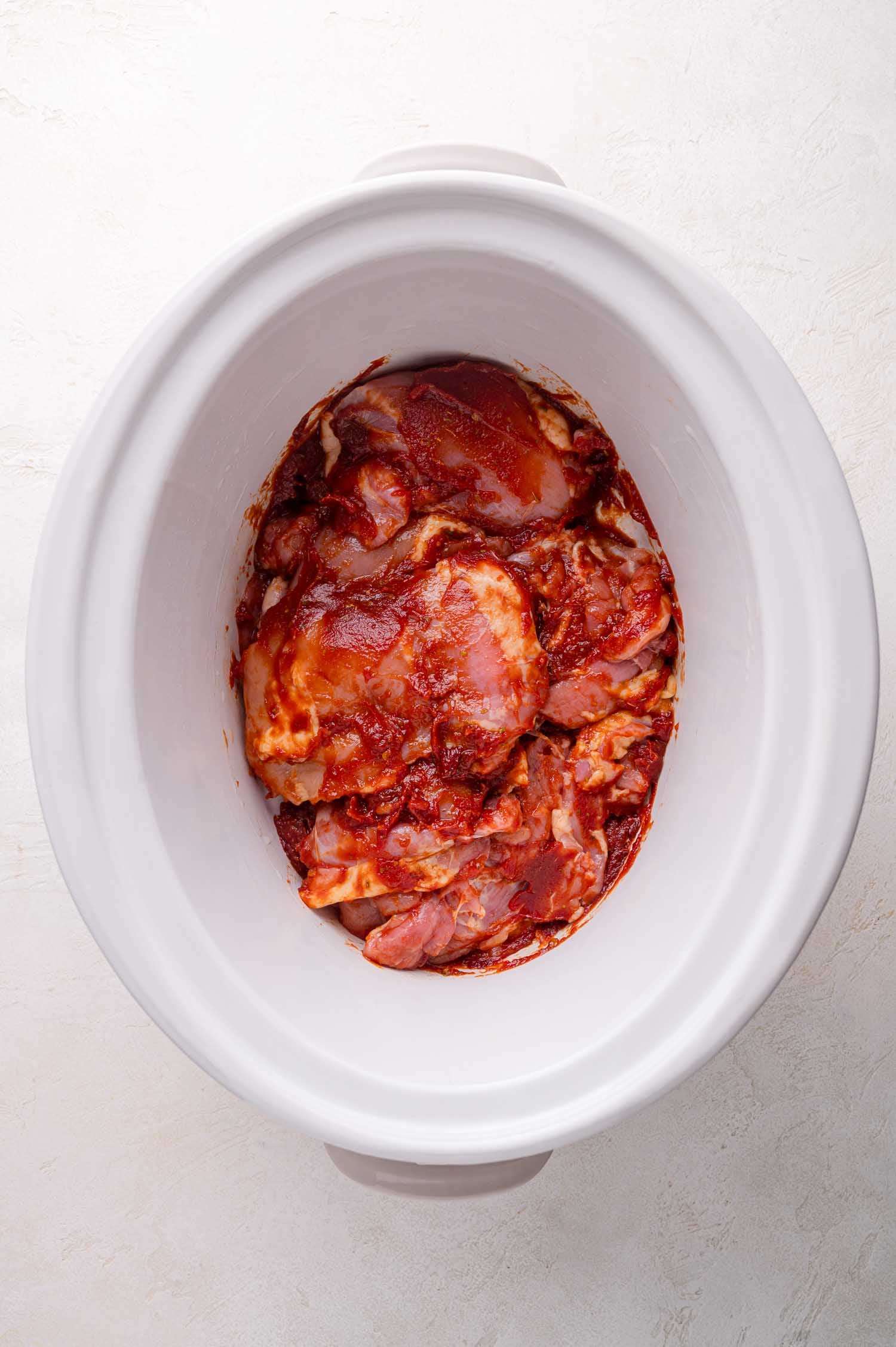 Raw, chicken thighs in a chipotle sauce a white slow cooker on a light-colored surface.