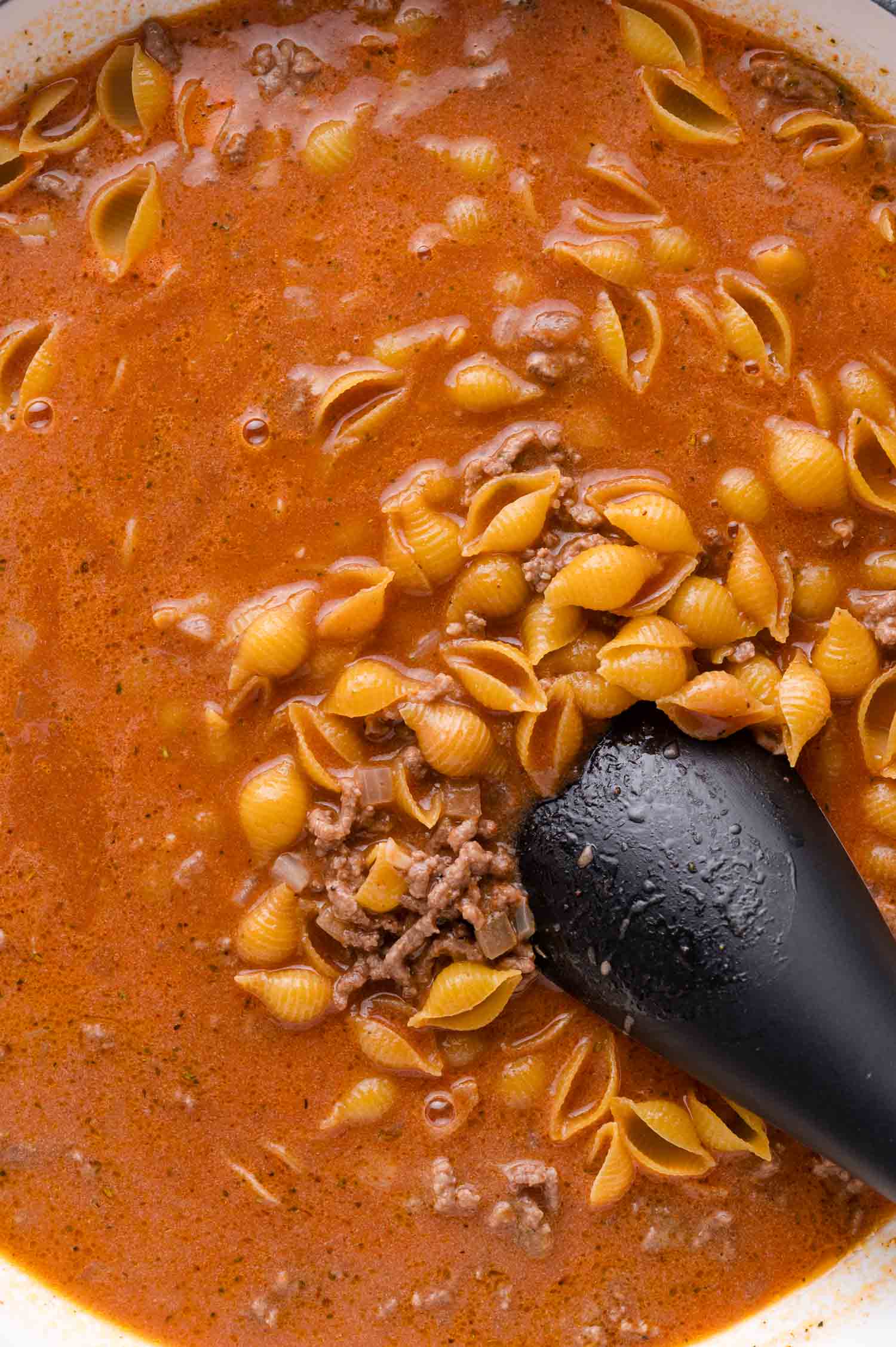 A close-up of a pot filled with shell pasta, ground beef, and tomato-based sauce being stirred with a black spoon.