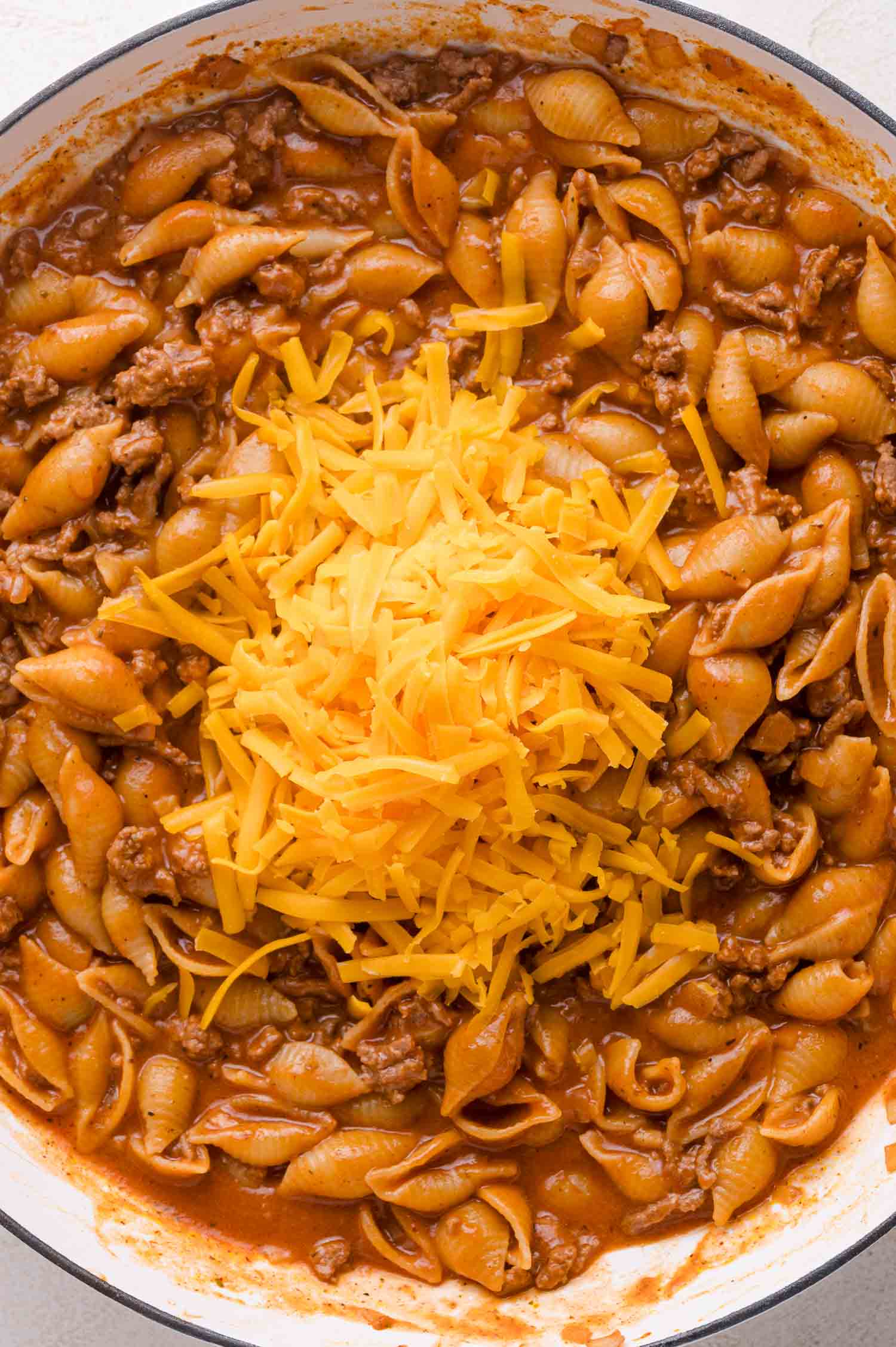 Cooked pasta shells with ground beef in a tomato-based sauce, topped with a mound of shredded cheddar cheese in the center of a pan.