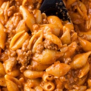 Close-up of cooked pasta shells mixed with ground beef and a creamy tomato sauce, being stirred with a black spoon.