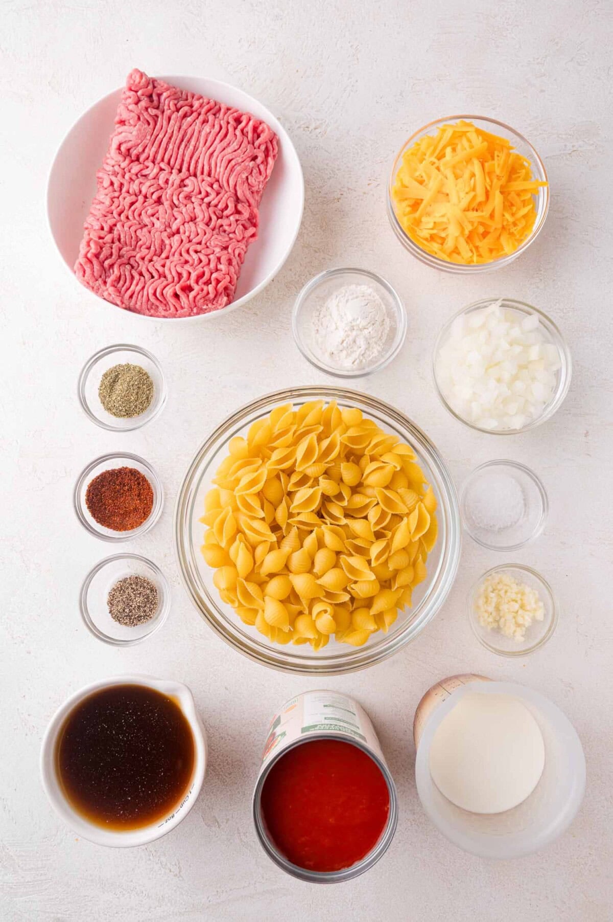 Ingredients for a pasta dish laid out on a white surface, including ground beef, pasta shells, shredded cheese, onion, spices, broth, tomato sauce, cream, flour, and minced garlic.