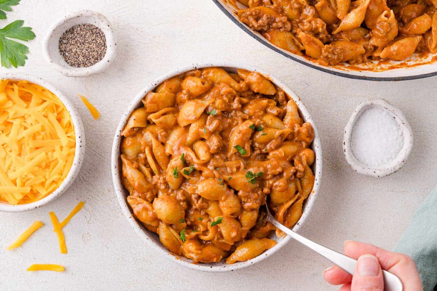 A bowl of pasta shells with ground meat in a creamy tomato sauce, topped with chopped herbs, next to bowls of shredded cheese, black pepper, and salt.