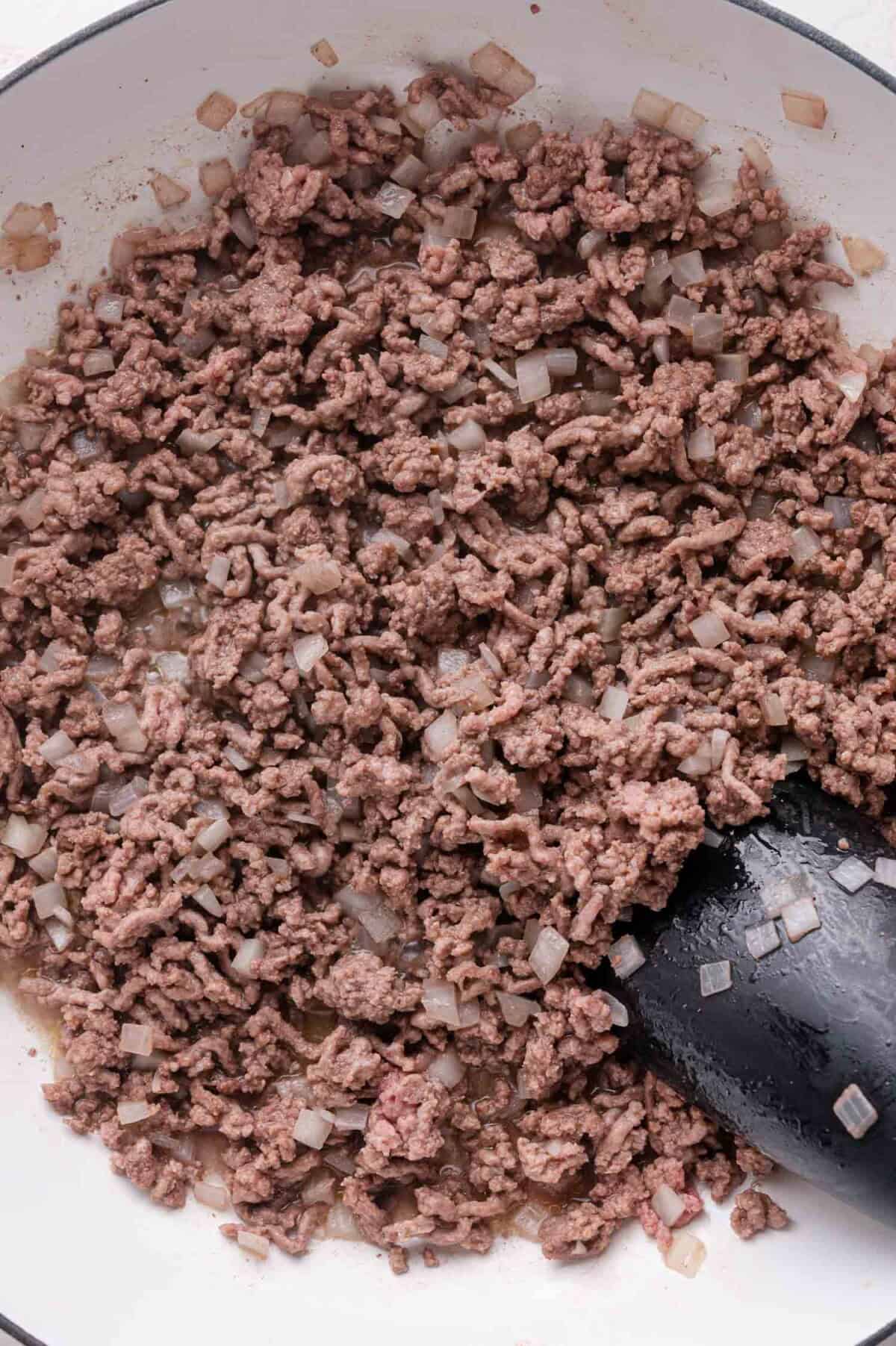 Ground beef and diced onions being browned in a white skillet with a black spatula.