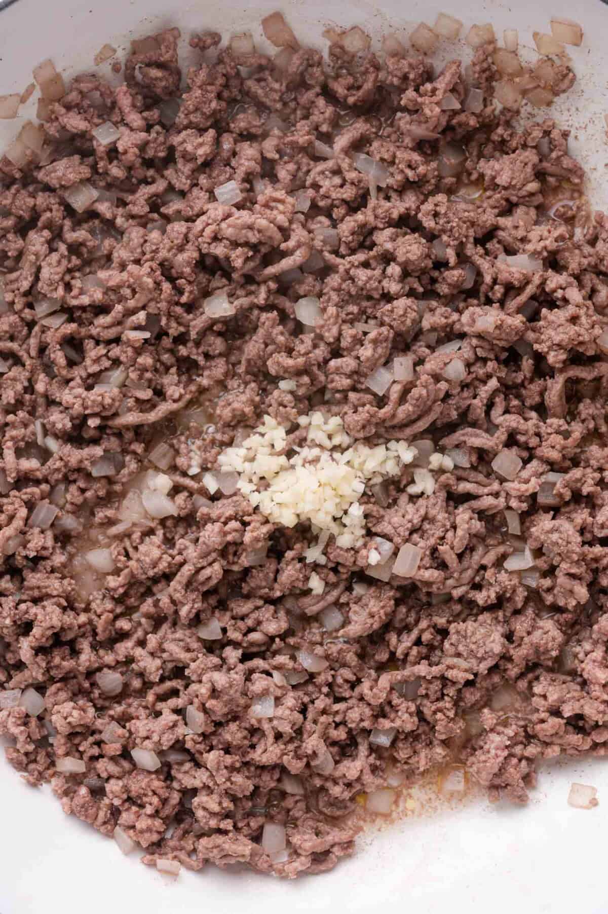 Ground beef and chopped onions cooking in a pan with a small pile of minced garlic added in the center.