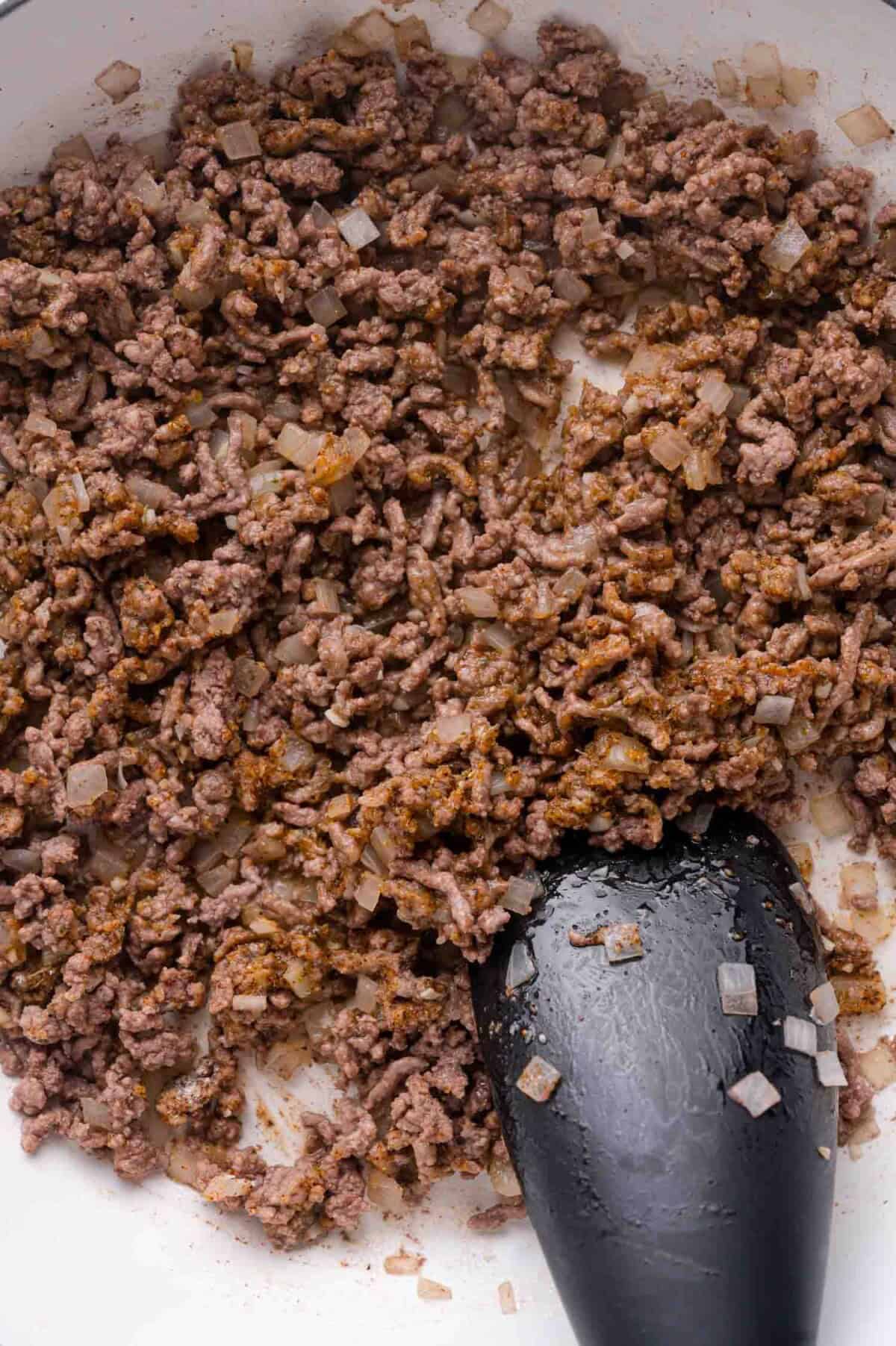 Ground beef and diced onions being cooked in a white pan with a black spatula.