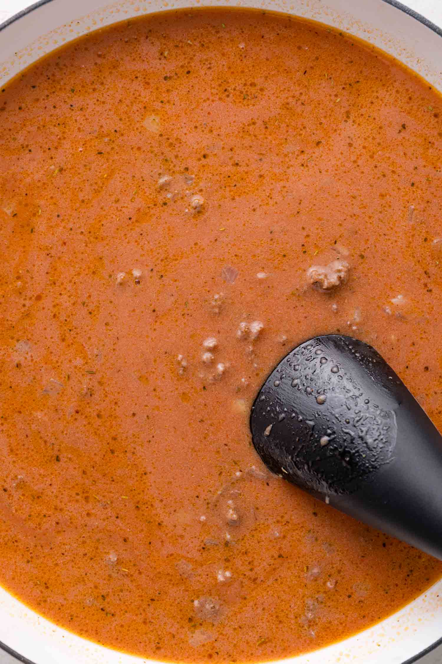 A close-up of a pot filled with creamy tomato sauce with visible ground meat, stirred with a black ladle.