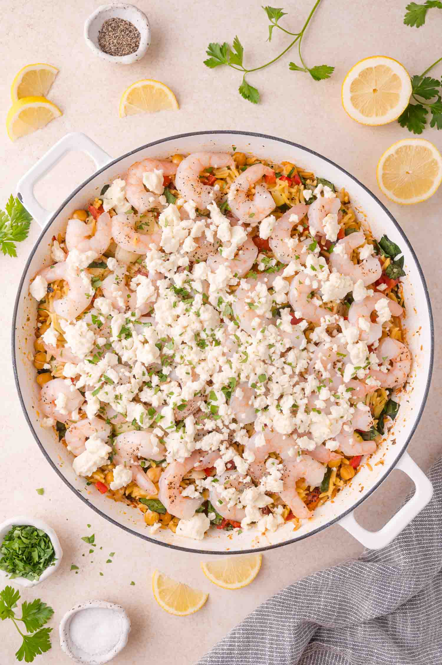A white pan filled with shrimp, crumbled feta cheese, vegetables, and orzo, surrounded by lemon slices, parsley, and small bowls of seasoning on a light countertop.