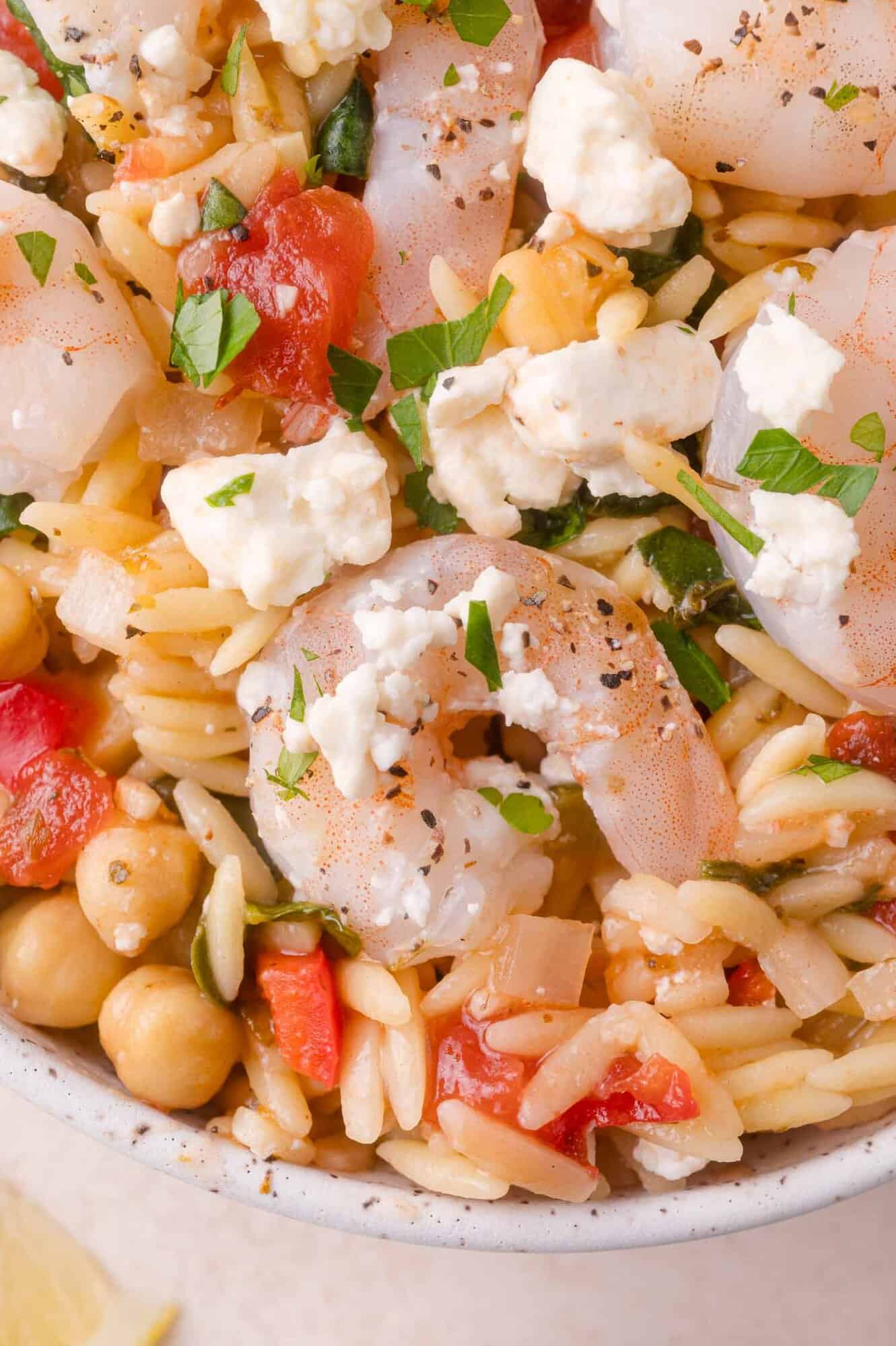 Close-up of a pasta salad with shrimp, chickpeas, crumbled feta cheese, diced tomatoes, herbs, and orzo in a white bowl.