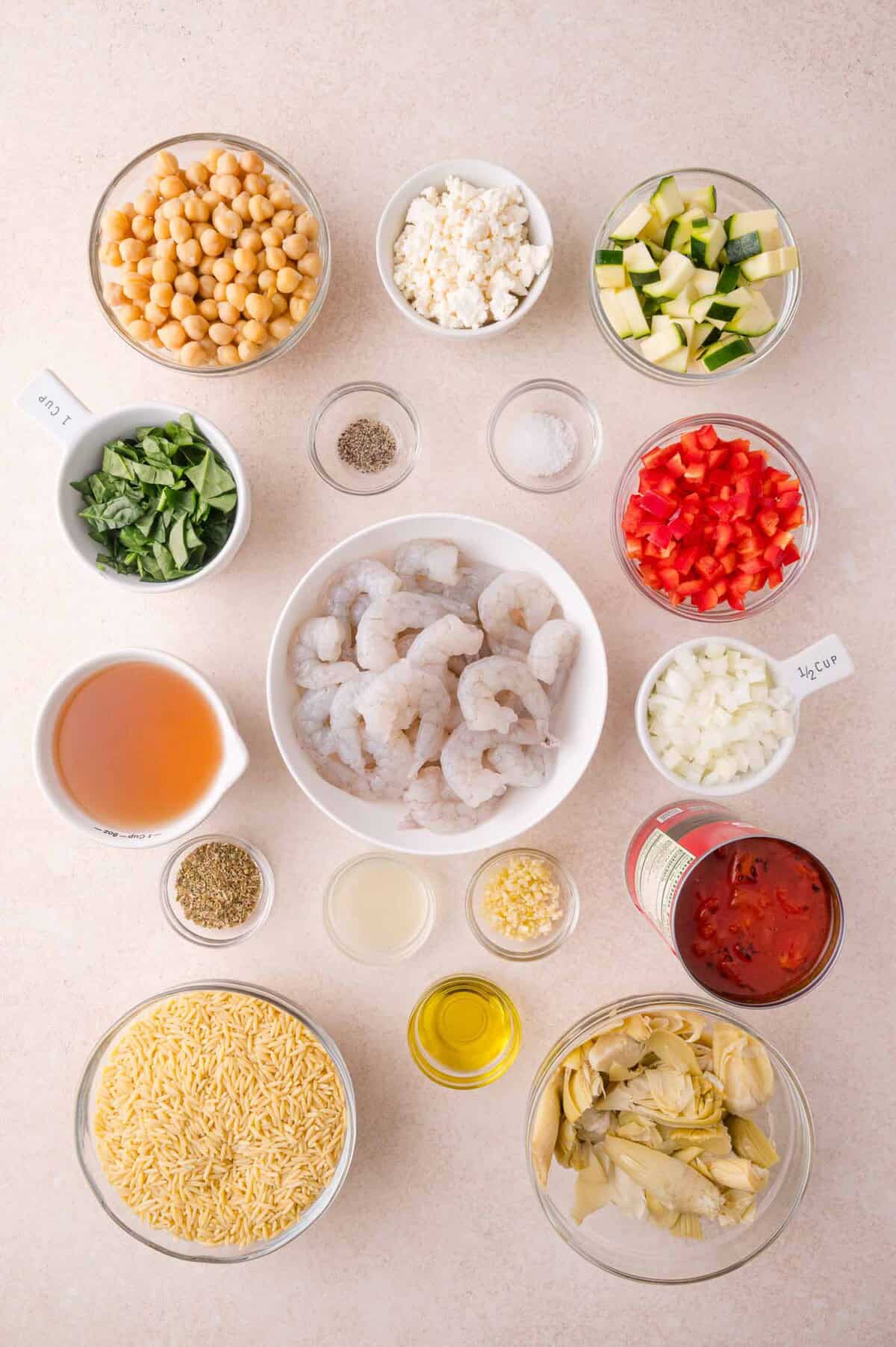 An overhead view of measured ingredients in bowls, including shrimp, orzo, chickpeas, artichokes, chopped vegetables, tomato, feta cheese, broth, herbs, oil, and seasonings on a light surface.
