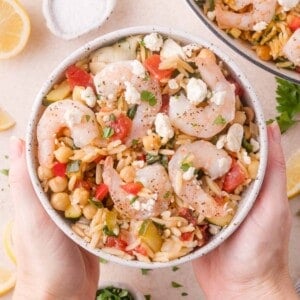 A bowl of shrimp orzo salad with chickpeas, tomatoes, zucchini, crumbled feta, and herbs, held by two hands.