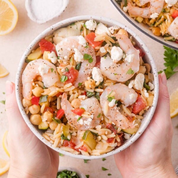 A bowl of shrimp orzo salad with chickpeas, tomatoes, zucchini, crumbled feta, and herbs, held by two hands.