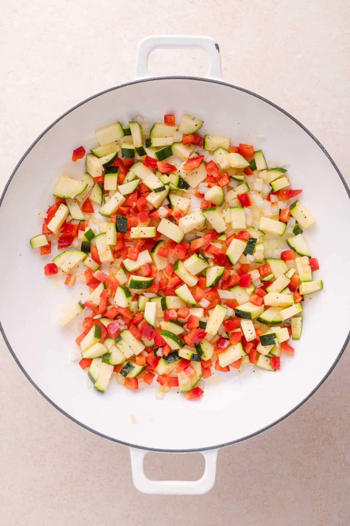 Diced zucchini, red bell pepper, and onion sautéing in a white pan on a light surface.