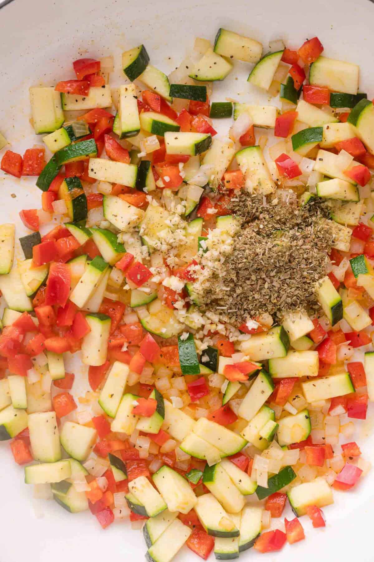 Chopped zucchini, red bell pepper, onion, minced garlic, and dried herbs sautéing in a pan.
