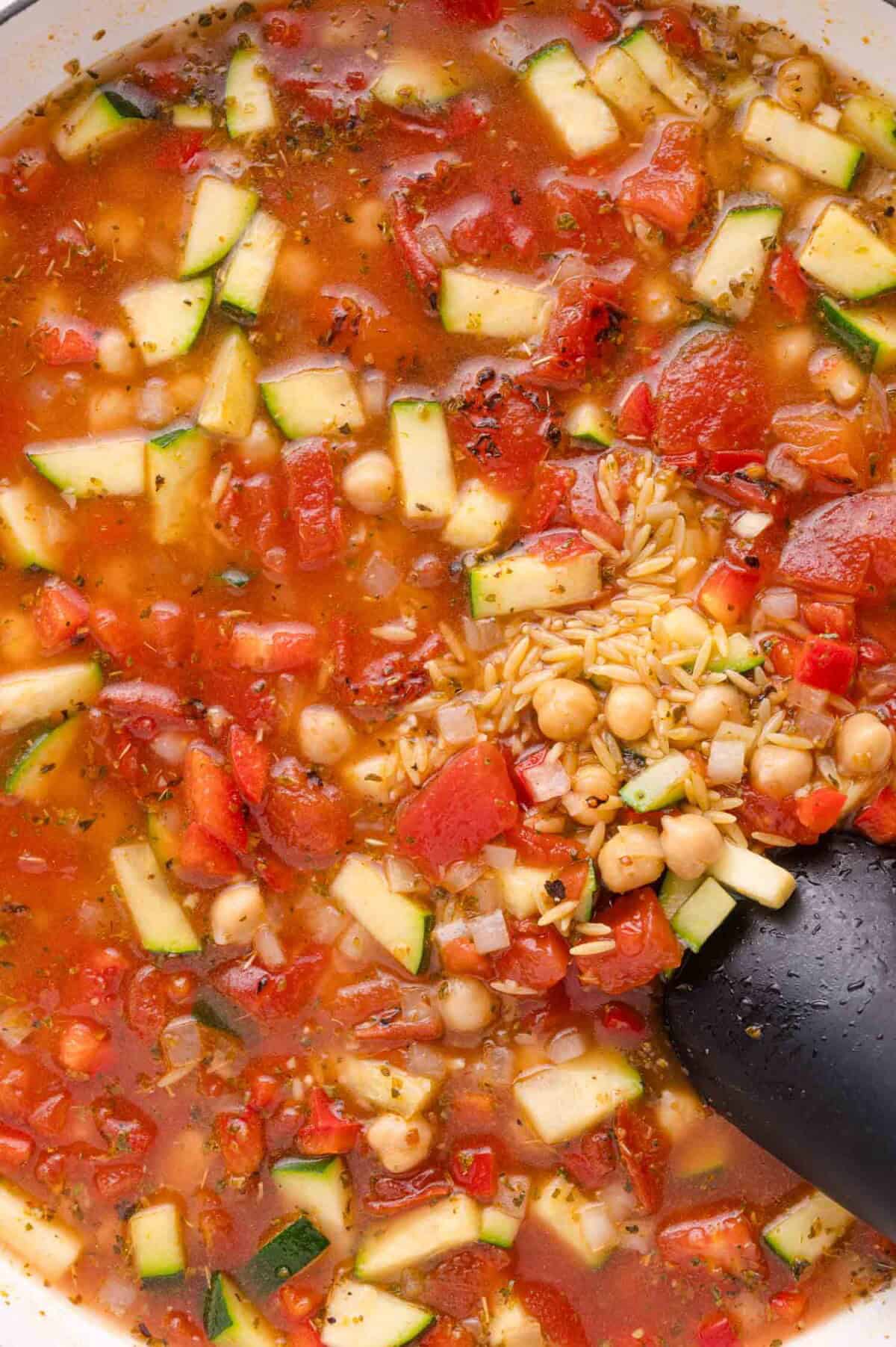 A pot of vegetable soup with chopped zucchini, chickpeas, tomatoes, herbs, and dry orzo pasta being stirred with a black spoon.