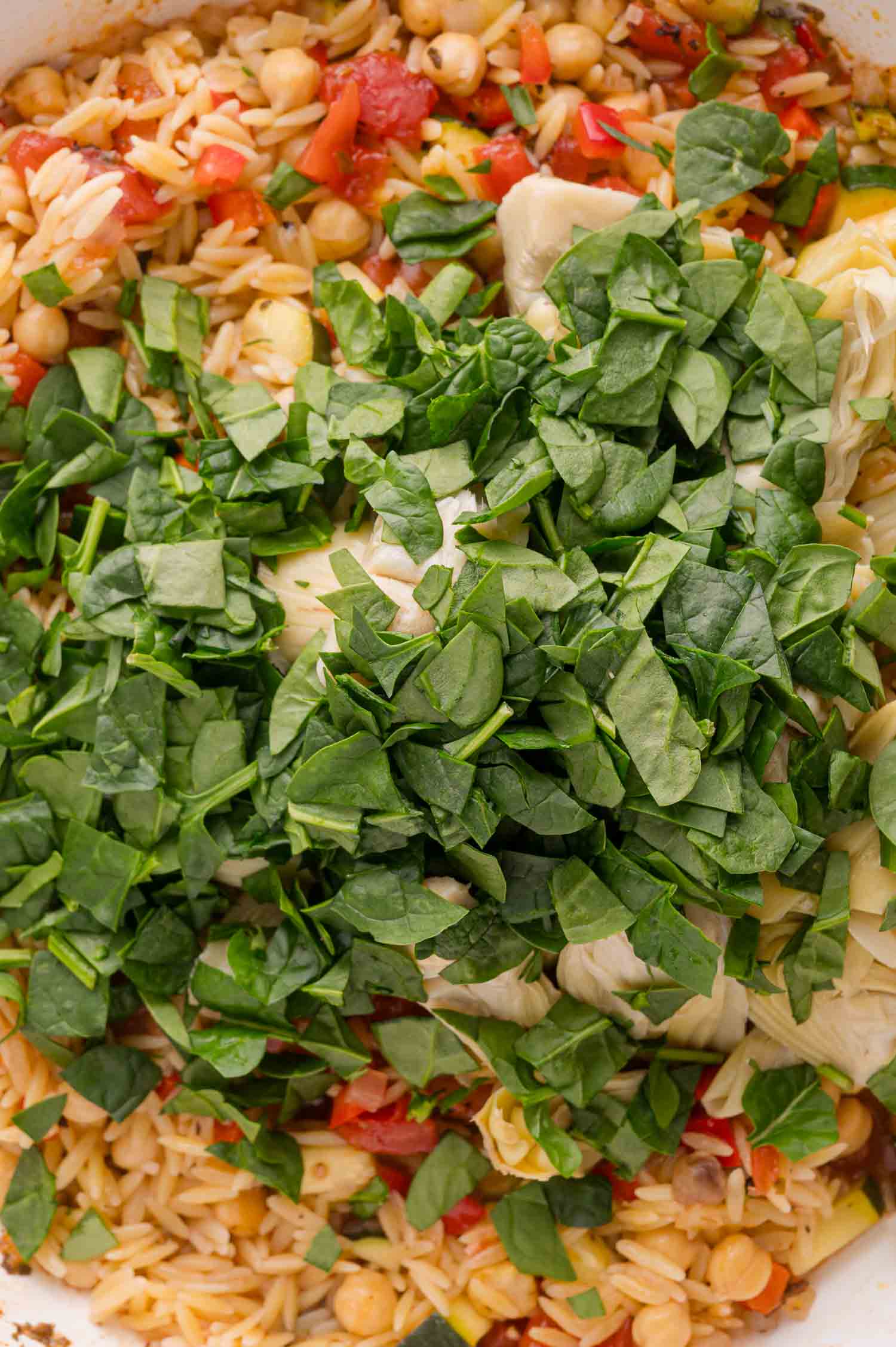 Chopped spinach on top of a mixture of orzo pasta, chickpeas, tomatoes, and artichokes in a pan.