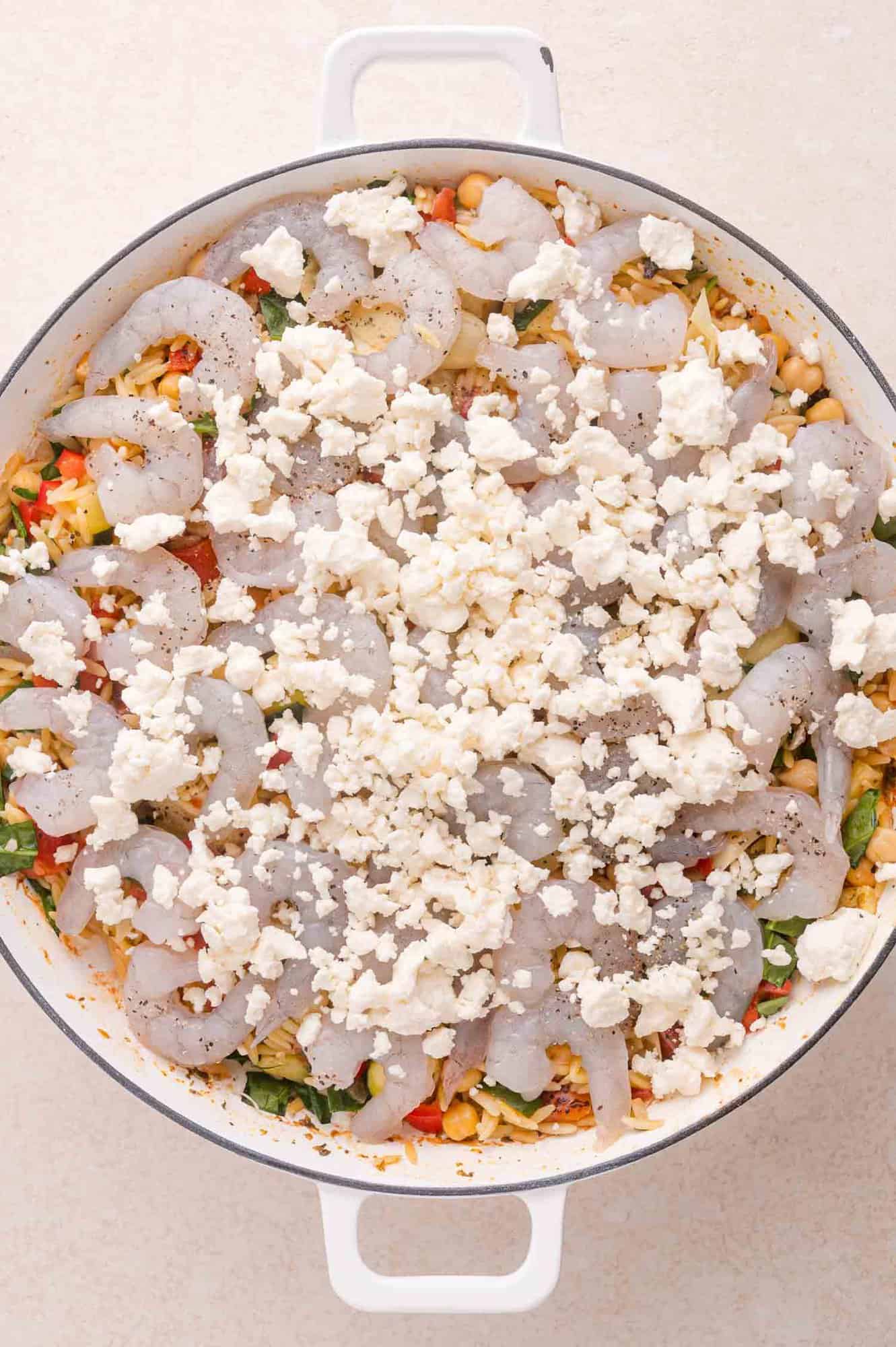 A white pan filled with uncooked shrimp, crumbled feta cheese, chopped vegetables, and chickpeas, ready to be cooked.